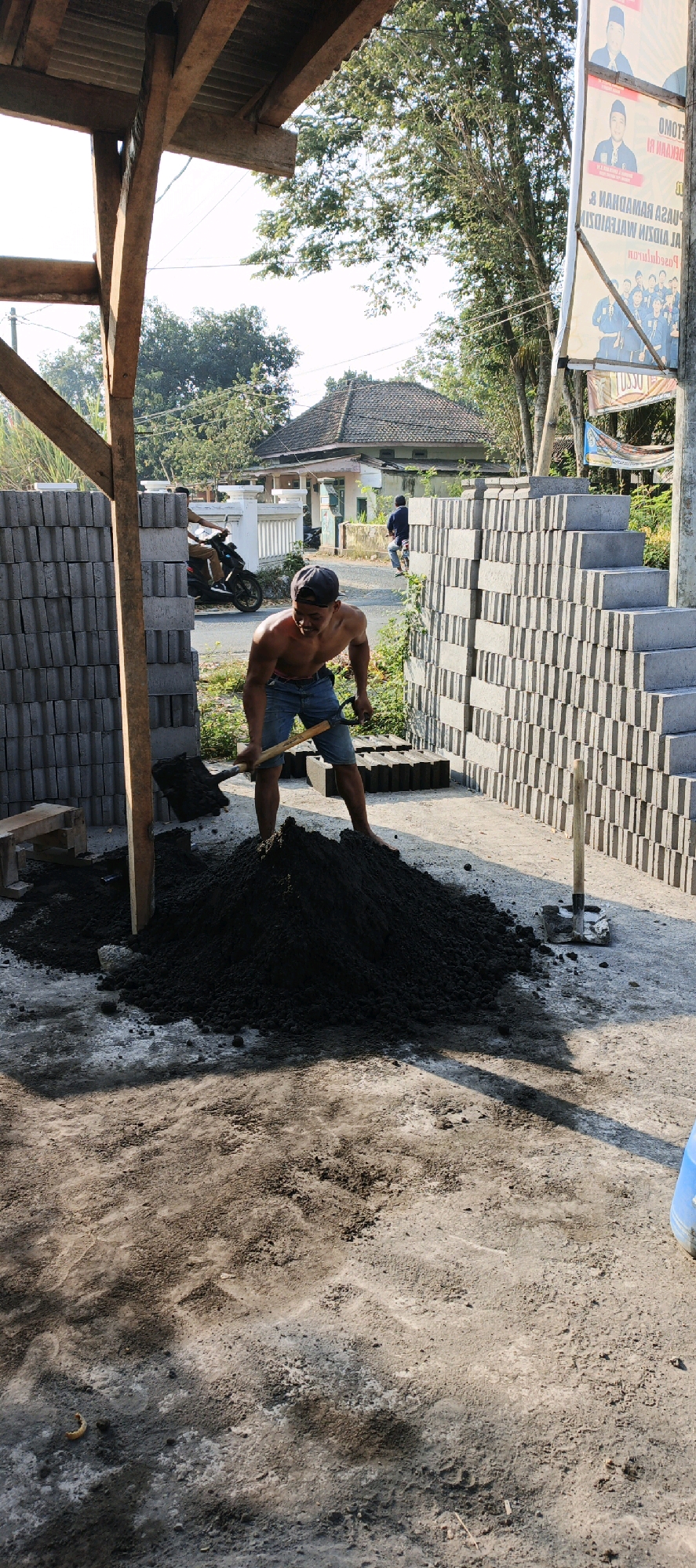 In Kasiyan, Indonesia, a shirtless young man is shoveling a pile of dark, granular material, likely cement or soil, on a dirt ground. He wears denim shorts and a dark baseball cap. To his left, stacks of gray concrete blocks are arranged. To his right, more concrete blocks are stacked, forming a wall. A wooden structure, possibly a partial roof support, stands to the left of the man. In the background, a house with a tiled roof is visible, as well as trees and other vegetation. A scooter is parked on a road behind the man, with a person on it. A banner with images and text in Indonesian hangs on a pole to the right of the scene. The overall environment appears to be an outdoor construction or material storage area under daylight.
