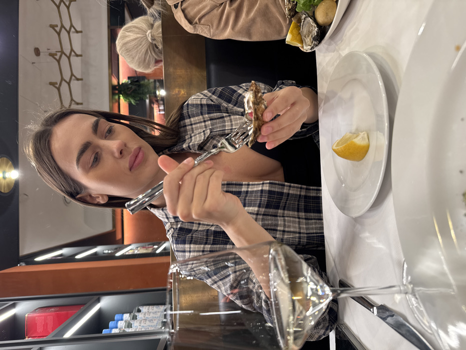 The image depicts an indoor dining scene within a modern restaurant located in Verona, Italy. A young adult female with long brown hair, wearing a blue and white plaid shirt over a dark top, is the central figure. She is seated at a table with a white tablecloth, holding an oyster in its half-shell with her left hand and a small silver fork in her right hand, positioned to eat the oyster. Her gaze is directed downwards towards the food. In the foreground to her left, a clear stemmed wine glass, partially filled with a pale yellow liquid, stands next to a silver knife. A small white plate with a lemon wedge is situated directly in front of her, and another white plate containing more oysters and lemon slices is visible to her right. The restaurant background features dark, possibly leather, booth seating, warm-toned orange walls, and shelving units with illuminated bottles and red boxes. A blonde-haired individual, facing away, is partially visible at a table behind the main subject. Overhead, a metallic, honeycomb-patterned light fixture and recessed ceiling lights are visible.