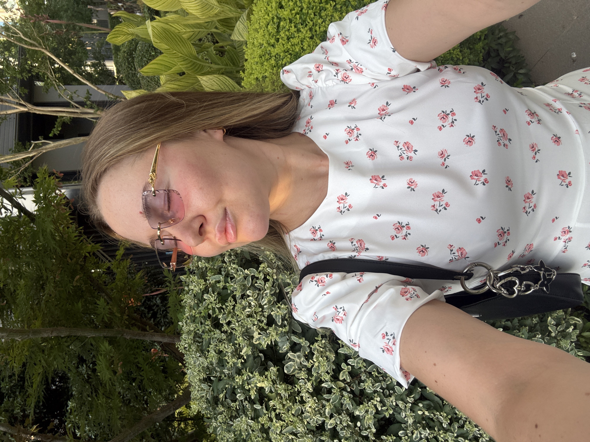 A young woman with blonde hair and pink-tinted sunglasses is taking a selfie outdoors. She is wearing a white dress with a floral print. Behind her is a lush green bush and some trees, suggesting an outdoor setting, possibly a park or garden. The lighting indicates it is daytime, likely late afternoon, as sunlight casts shadows. There is no indication of this location being in Yiwu, China.
