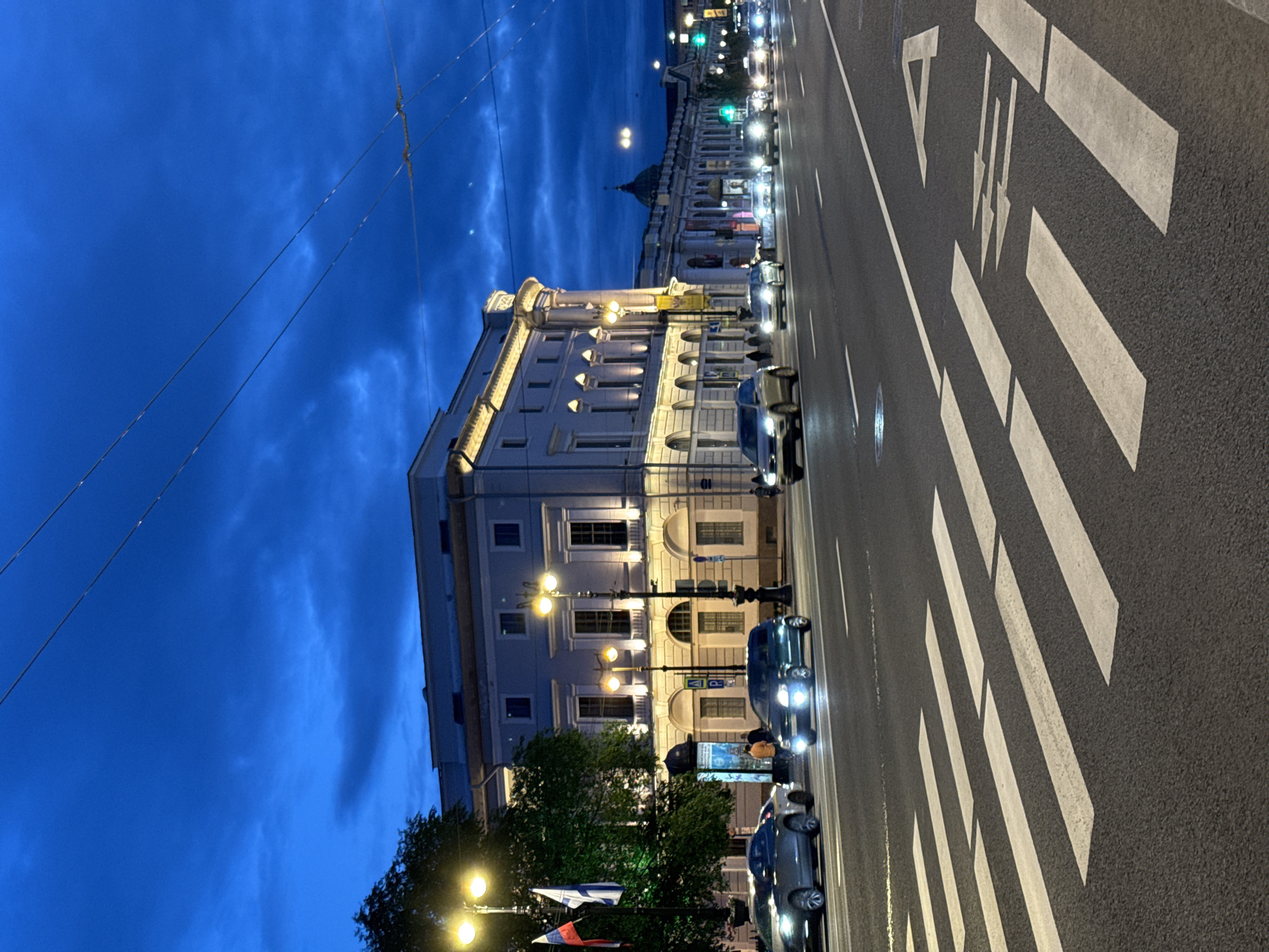 This is a medium shot, taken at dusk or night, of a street scene in Saint Petersburg, Russia, likely Nevsky Prospekt, given the architecture. A wide, multi-lane road dominates the foreground, marked with white pedestrian crossing stripes and directional arrows. Several cars are driving on the road, their headlights illuminated. On the left side of the road, a grand, light-colored building with classical architectural features stands illuminated by streetlights. Flags are visible on poles in front of the building. On the right side of the road, a long, continuous building with arched windows stretches into the distance, also illuminated. The sky above is a deep, dark blue, with dramatic clouds and visible power lines.