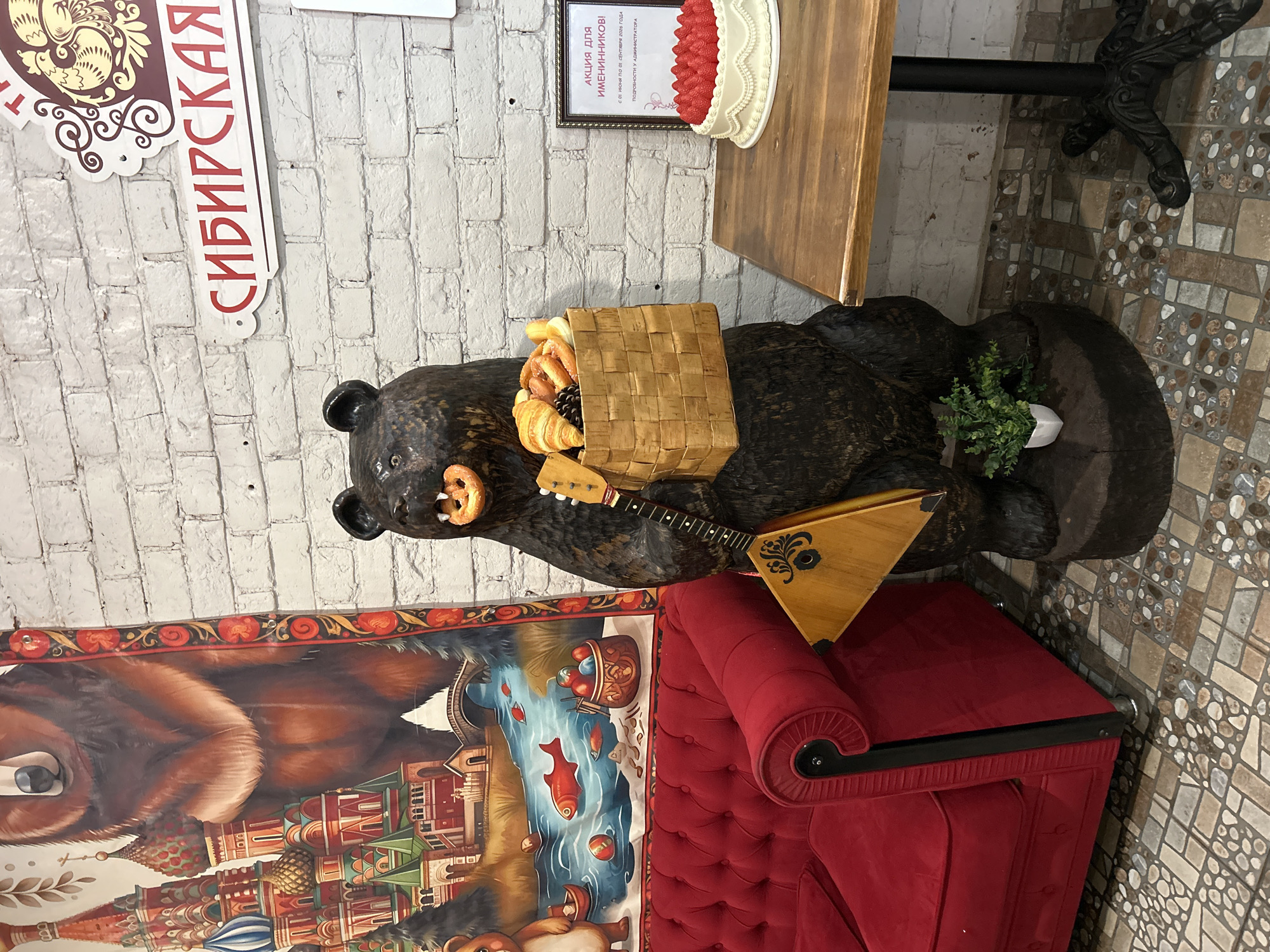 The image captures a scene indoors, likely within a establishment in Tomsk, Russia, as indicated by a sign that reads "СИБИРСКАЯ" (Sibirskaya, meaning Siberian). The focal point is a life-sized wooden sculpture of a bear, intricately carved and painted. The bear is depicted holding a woven basket filled with various baked goods, including croissants, pretzels, and what appear to be small buns. In its mouth, the bear has a pretzel. It is also holding a balalaika, a traditional Russian stringed instrument, in its left arm.
To the left of the bear sculpture is a plush red sofa with tufted upholstery, suggesting a seating area for customers. Behind the sofa and the bear is a large, colorful mural depicting a bear and iconic Russian architecture, possibly St. Basil's Cathedral, set against a backdrop of a river with fish.
To the right of the bear sculpture, a wooden table stands with a black, ornate pedestal base. On the table, a framed certificate is displayed, advertising a promotion for "АКЦИЯ ДЛЯ ИМЕНИННИКОВ!" (Aktsiya dlya imieninnikov!, meaning "Promotion for Birthday People!"). Below the certificate is a white cake decorated with a generous topping of fresh strawberries and white icing details.
The background consists of a white brick wall, adding a rustic element to the decor. The floor is tiled with a mosaic pattern of small, irregularly shaped stones in various earthy tones. A small potted green plant sits at the base of the bear sculpture, adding a touch of natural greenery to the scene. The lighting is ambient, illuminating the subjects without harsh shadows. There are no people visible in the image.