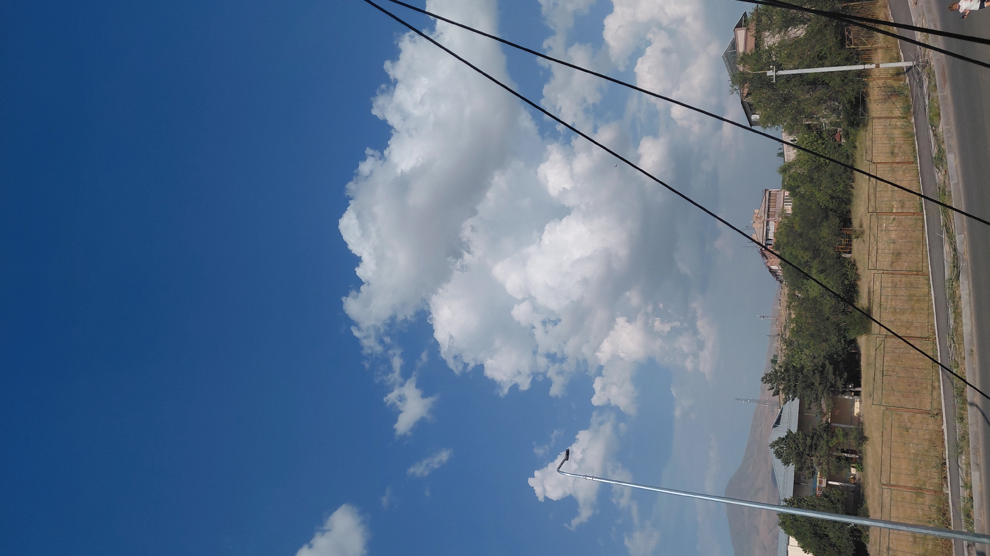 The image captures a street-level view in Abovyan, Armenia, under a bright blue sky dotted with fluffy white clouds. In the foreground, a section of an asphalt road is visible, with a paved sidewalk adjacent to it. A rusty metal fence runs along the edge of the sidewalk, separating it from a dry, grassy area with scattered trees. Behind the fence, a building with a light-colored exterior and a dark roof is partially visible. Further back, more trees and buildings are scattered across the landscape. A tall, silver lamppost stands on the left side of the image, and several black power lines stretch diagonally across the frame from the right. The sky is a prominent feature, with a large cluster of cumulus clouds dominating the upper portion of the image. A mountain is visible in the distance on the left, shrouded in haze.