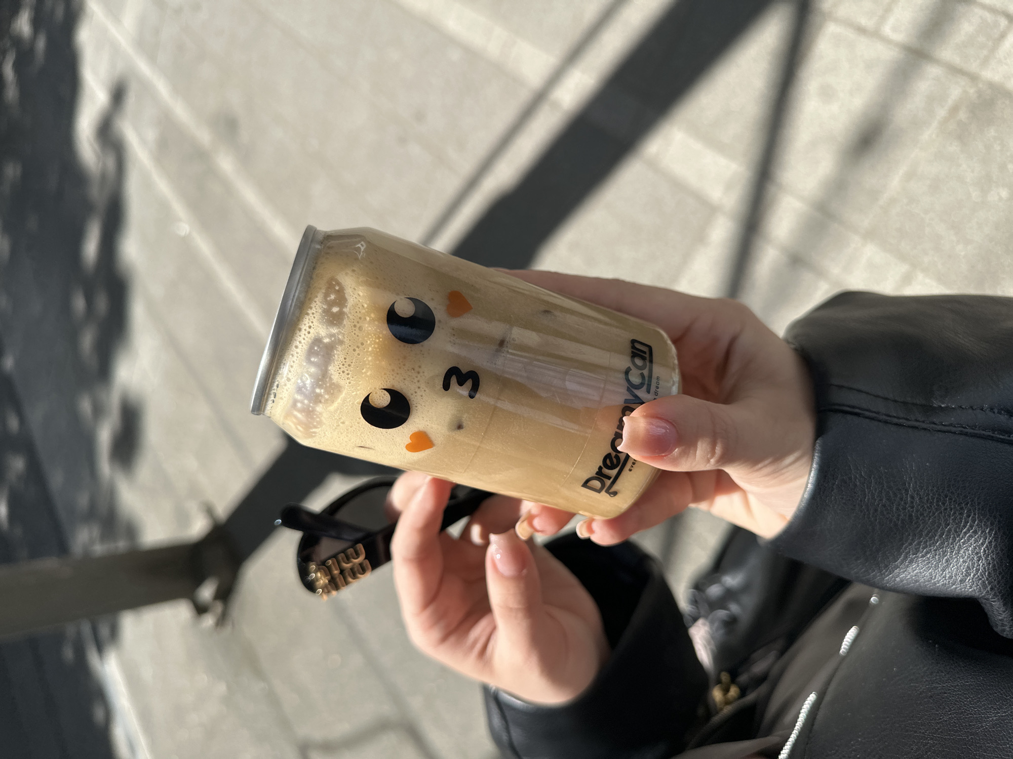 A person wearing a black leather jacket holds a can of DreamyCan iced coffee. The can features a cartoon face. Sunglasses are also visible in the person's other hand. The setting appears to be outdoors on a paved surface, possibly in Belgrade, Serbia. Light and shadows suggest it is daytime.