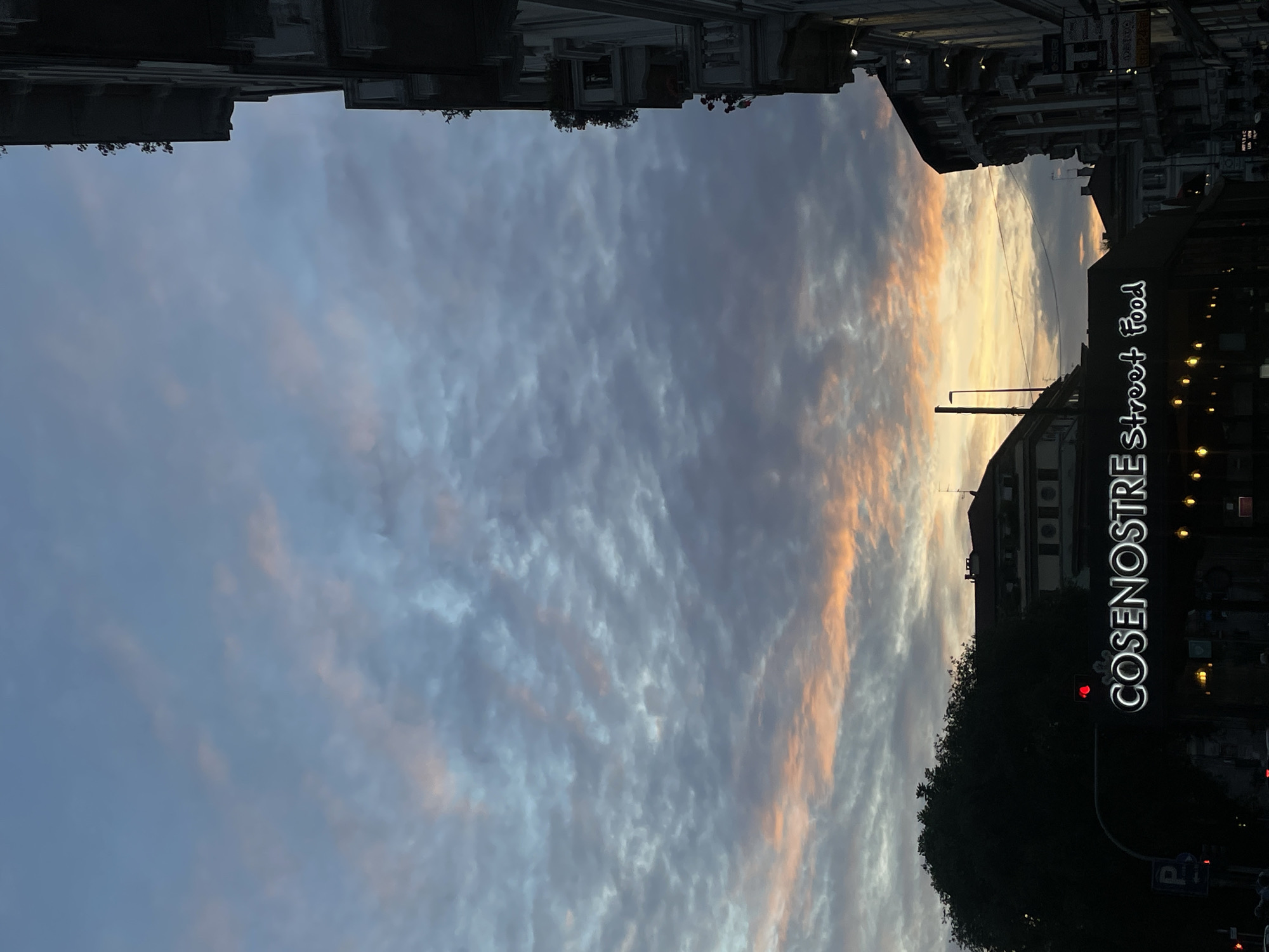 The image depicts a street in Milan, Italy, at dusk. The sky is filled with layered clouds, some tinged with orange hues from the setting sun. Buildings line the street, with "COSENOSTRE Street Food" illuminated in bright white letters on one side, possibly a storefront sign. Trees are visible, silhouetted against the evening sky. A traffic light is visible, displaying a red signal. The scene suggests a city environment at twilight.