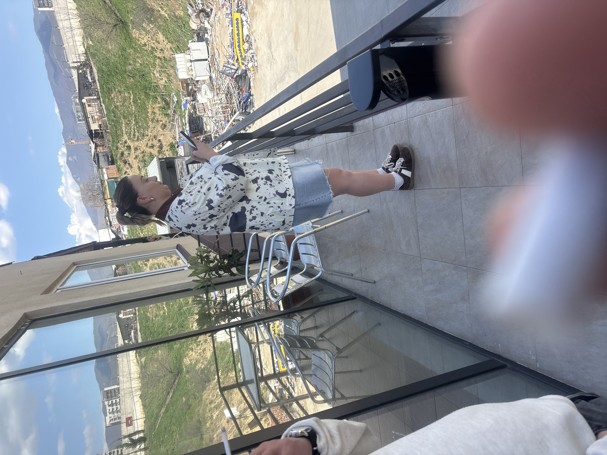 On a balcony overlooking a cityscape in Tbilisi, Georgia, a woman stands looking at her smartphone. She wears a white jacket with a black print, a denim skirt, white socks, and brown shoes. The balcony is tiled and furnished with metal chairs. A black barbecue grill is visible. Outside the balcony, the landscape includes greenery, mountains, buildings, and a junkyard-like area. A building with large windows reflects the sky and landscape. In the foreground, parts of a person and a lit cigarette are visible. The lighting suggests it's daytime. The sky is blue with some clouds.