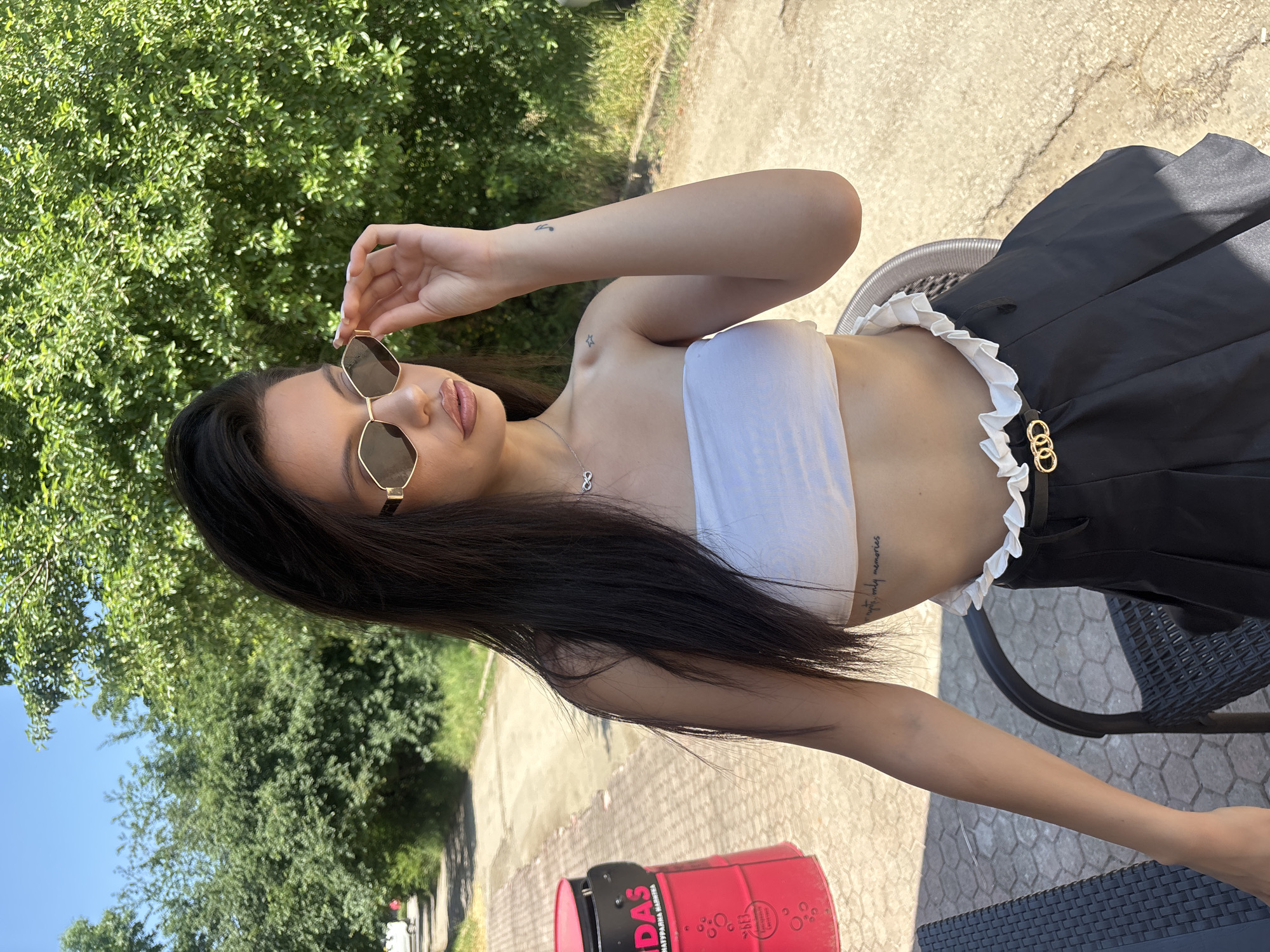 A young woman with dark hair, wearing sunglasses, a white tube top, and a black pleated skirt with a ruffled waistband, is seated outdoors in Sofia, Bulgaria. She is holding her sunglasses with her right hand. The background features green trees, a pathway, and a red cylindrical object with text. The ground is paved with hexagonal tiles. The lighting suggests it is daytime, likely sunny.