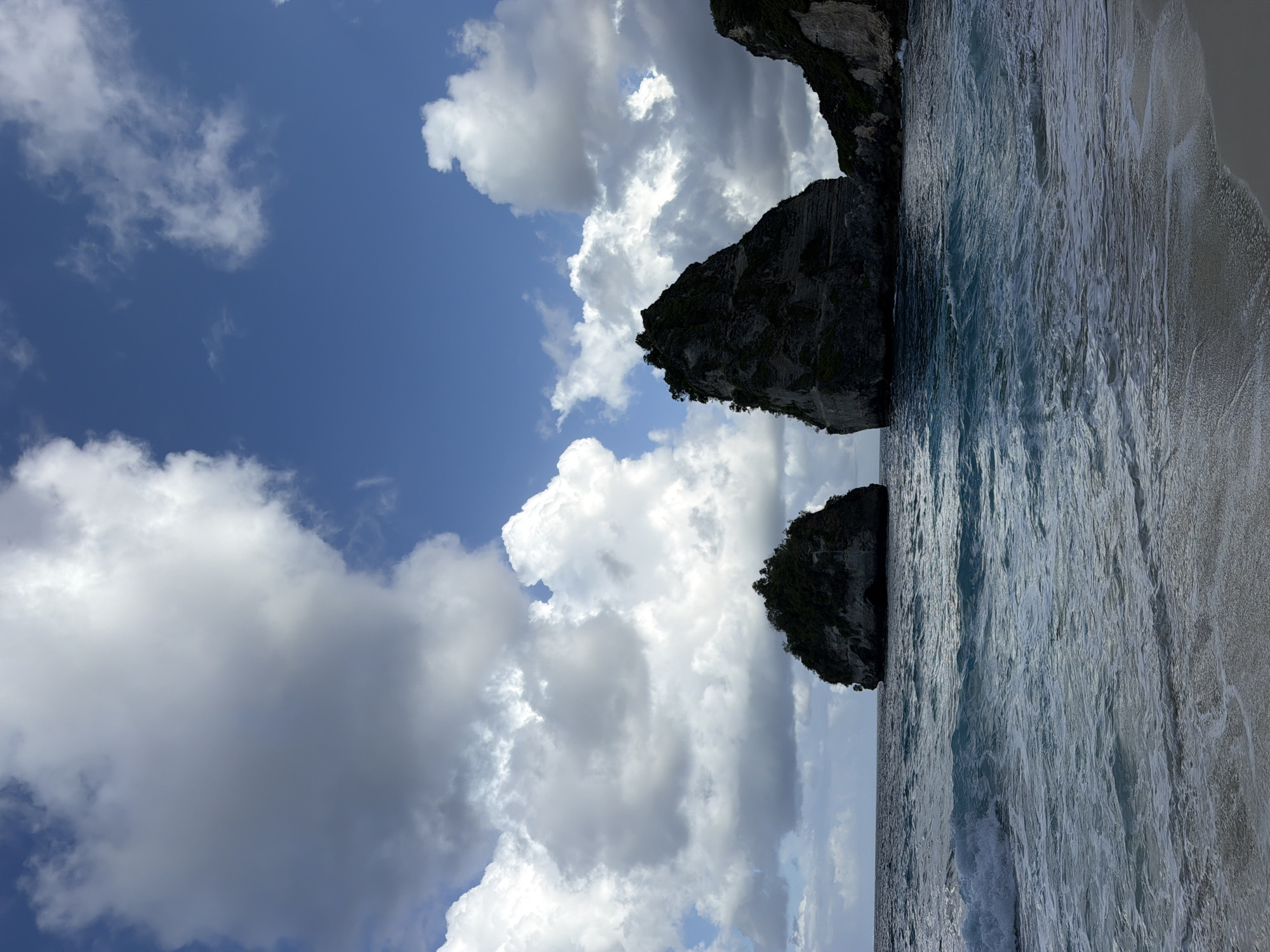 This is a landscape photograph of a tropical beach with dramatic rock formations jutting out of the ocean. The foreground shows a sandy beach with gentle waves lapping at the shore. The water is a clear turquoise, transitioning to a deeper blue further out. Two prominent rock formations, densely vegetated with trees, are visible in the mid-ground. The larger formation on the right is a towering, jagged peak, while a smaller, rounder formation sits to its left. The background features a vast blue sky filled with large, fluffy white and grey clouds, creating a dynamic contrast with the ocean and rock structures. The scene is consistent with the iconic views found in Nusa Penida, Indonesia, specifically locations like Diamond Beach or Kelingking Beach, known for their dramatic cliffs and offshore rock formations. There are no people or man-made objects visible in the image.
