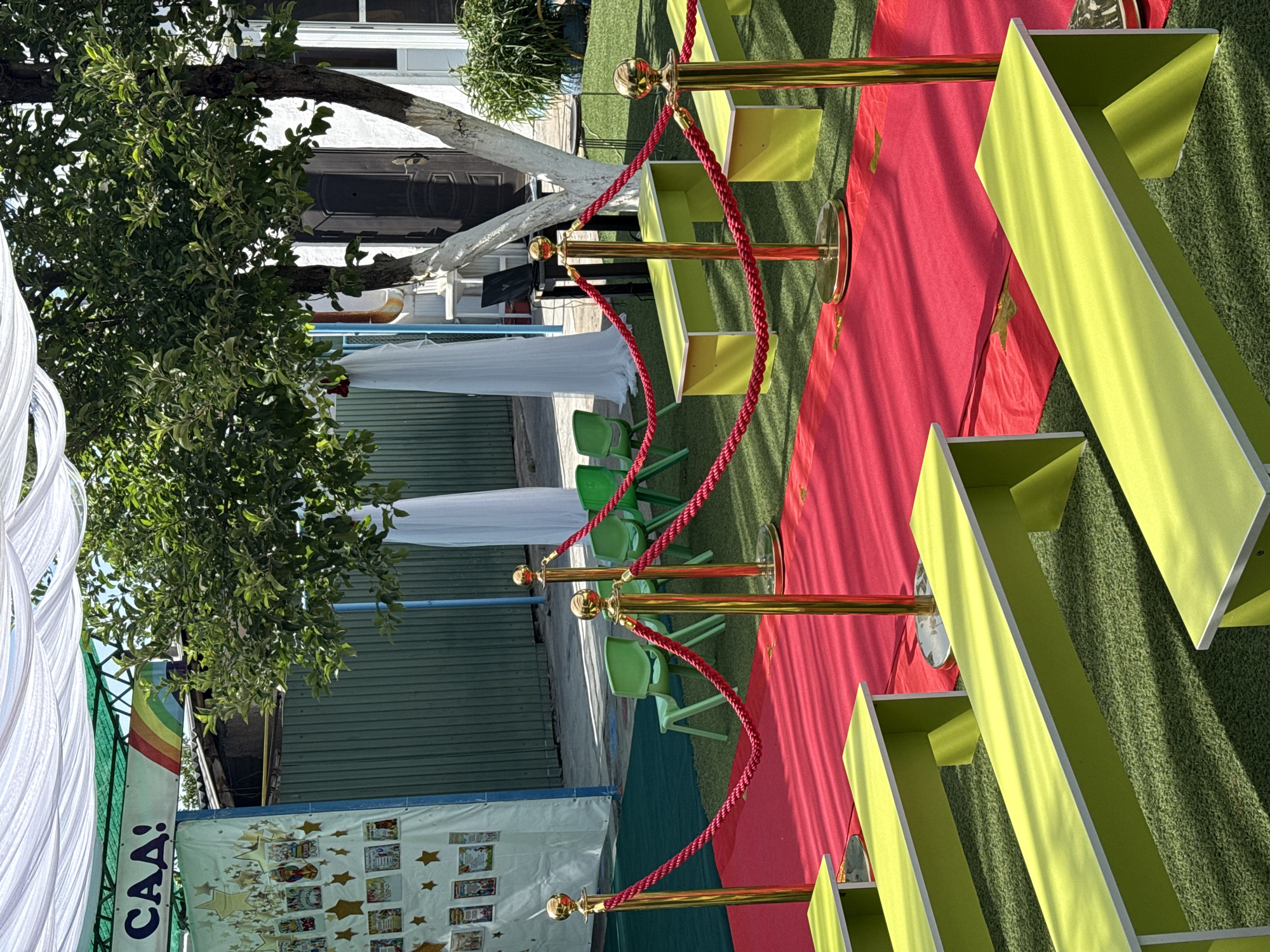 The image captures an outdoor scene in the city of Unknown, Unknown. A red carpet extends across a patch of artificial green turf, bordered by gold-colored stanchions linked with red ropes. The stanchions guide the flow of pedestrian traffic. To either side of the carpet are rectangular, open-faced shelving units painted a light green color.
In the background, one can see several lime-green plastic chairs. A large tree provides shade with its dense canopy of green leaves. A white fabric canopy is also visible, along with a sign featuring a rainbow and the letters "CAA" above various images, possibly diplomas or children's artwork. A building with a painted white tree trunk and a dark door is visible in the background, adding depth to the composition.