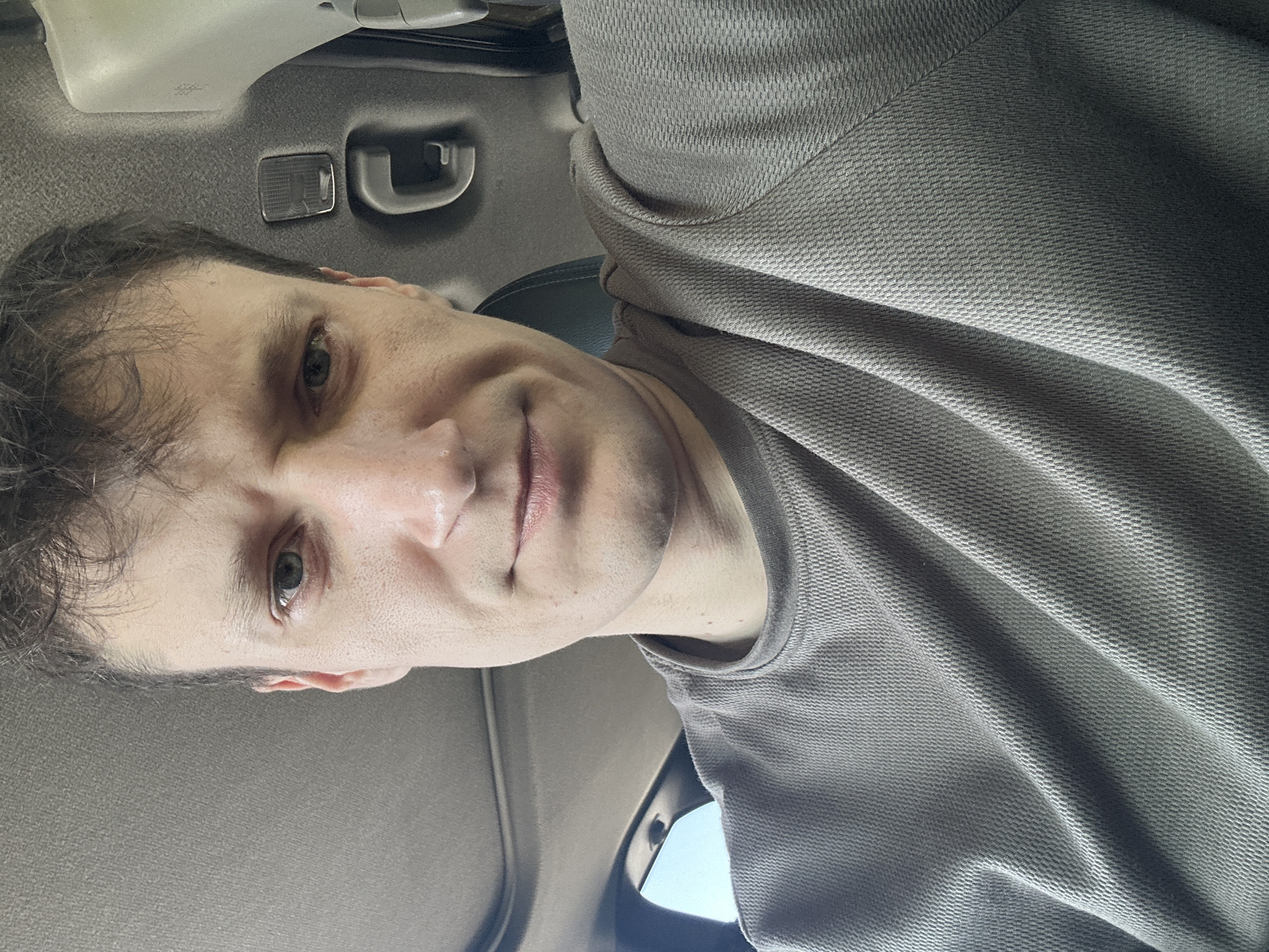 A close-up, selfie-style shot of a man's face and upper torso, taken from a low angle inside what appears to be a vehicle. The man has fair skin, light brown hair, and blue-green eyes. He is wearing a grey, short-sleeved, textured t-shirt. The background shows the interior of a car, with the headliner, a portion of a sunroof, a grab handle, and a light fixture visible. The lighting suggests it is daytime, likely outdoors. The image does not provide any information about the location in Makiv, Ukraine.