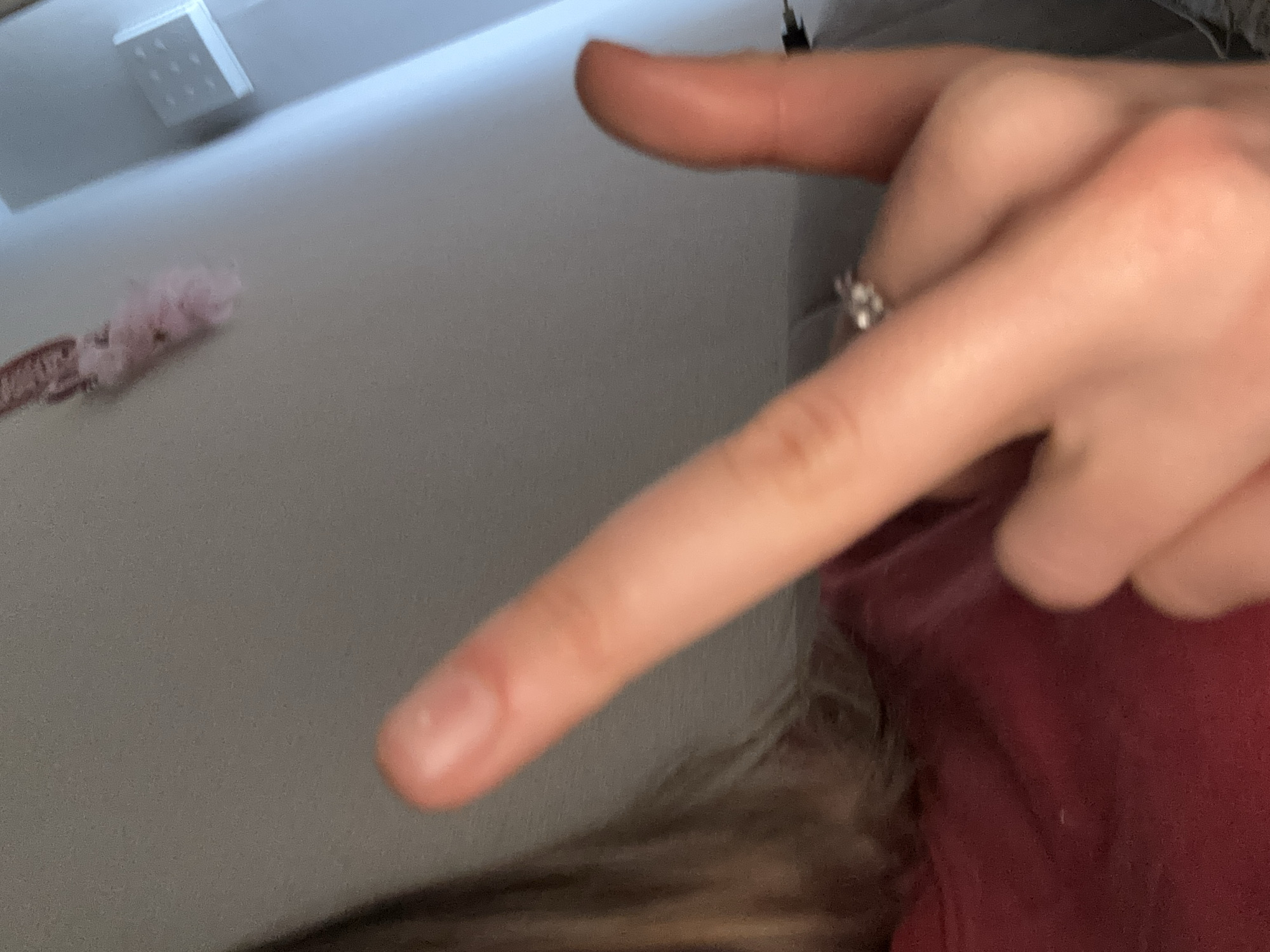 In the foreground, a person's right hand is shown, with the index finger and thumb extended in a gesture. A ring is visible on the ring finger. The person is wearing a dark red shirt. The background is blurred and appears to be an indoor setting with a wall and some objects. There is no information in the image that allows for the determination of the specific location within Vsevolozhsk, Russia.