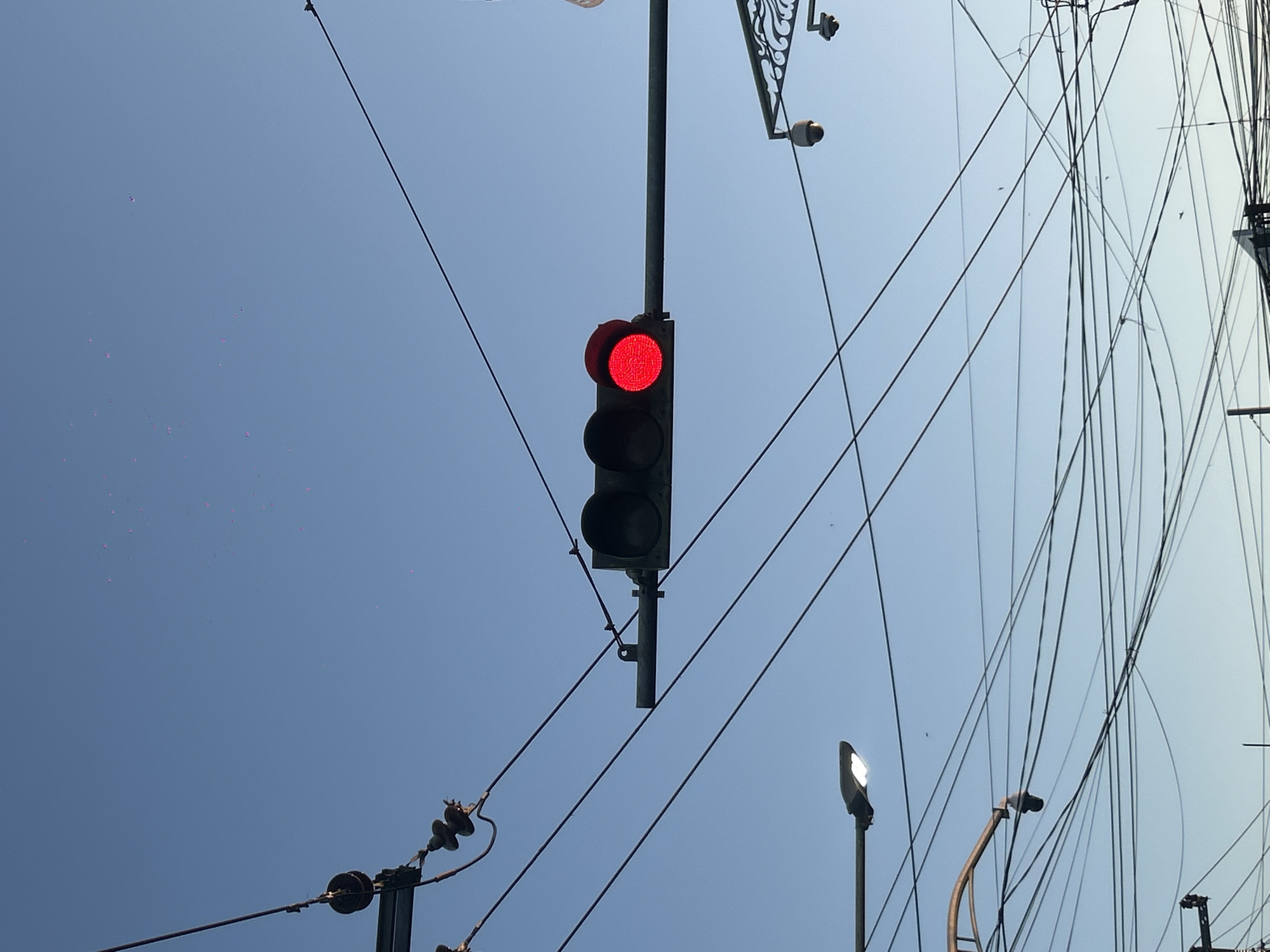 The image shows a traffic light in North Kuta, Indonesia, with the red light illuminated. The traffic light is mounted on a pole and surrounded by a network of overhead wires. The sky is clear and blue. Other utility poles with street lights are also visible. There are no people visible.