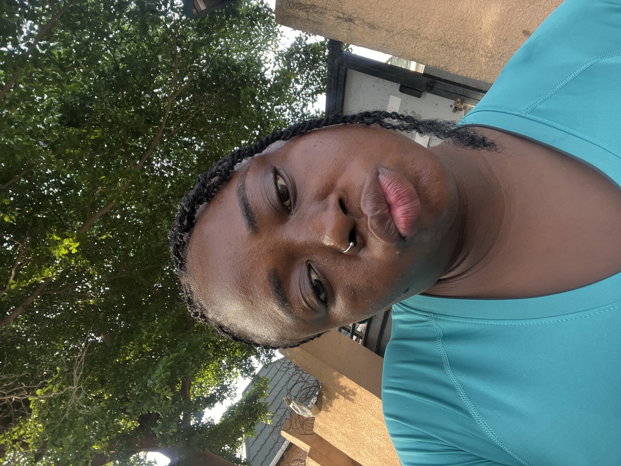 A selfie taken outdoors shows a woman with dark skin and braided hair, wearing a teal t-shirt. She has a nose ring and her lips are pursed. In the background, there are trees with green leaves, a fence with barbed wire, and a building. The overall setting appears to be urban, potentially in Abuja, Nigeria, given the architectural style of the fence and building. The lighting suggests it is daytime.