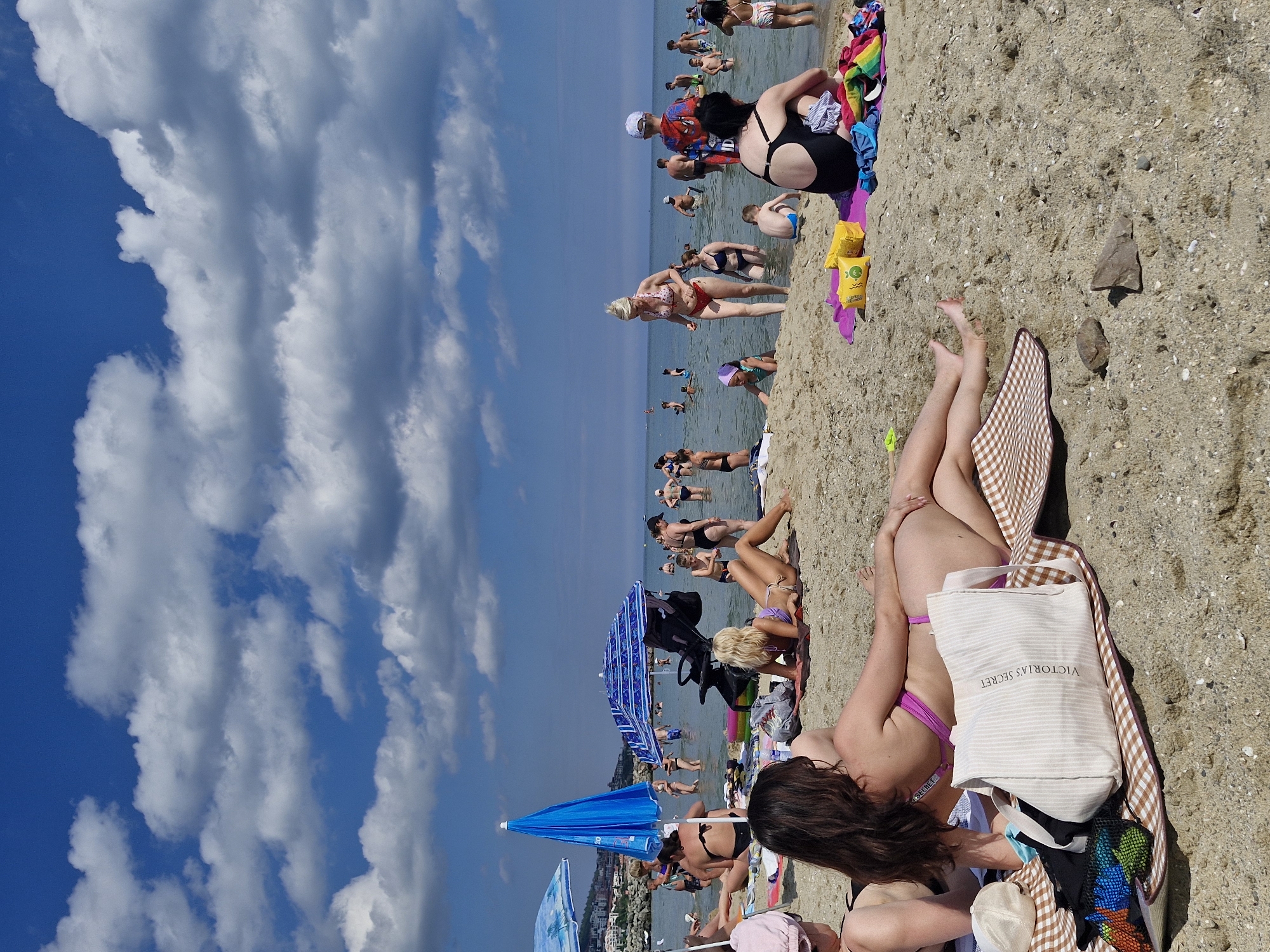 The image depicts a crowded beach scene in Sveti Vlas, Bulgaria, under a blue sky with scattered white clouds. Numerous people are visible, some bathing in the shallow sea, while others relax on the sandy shore. Several beach umbrellas provide shade. In the foreground, a woman is lying on a checkered blanket, with a large canvas bag labeled "VICTORIA'S SECRET" beside her. Other beach accessories, such as towels and bags, are scattered around. The overall atmosphere suggests a lively summer day at the seaside.