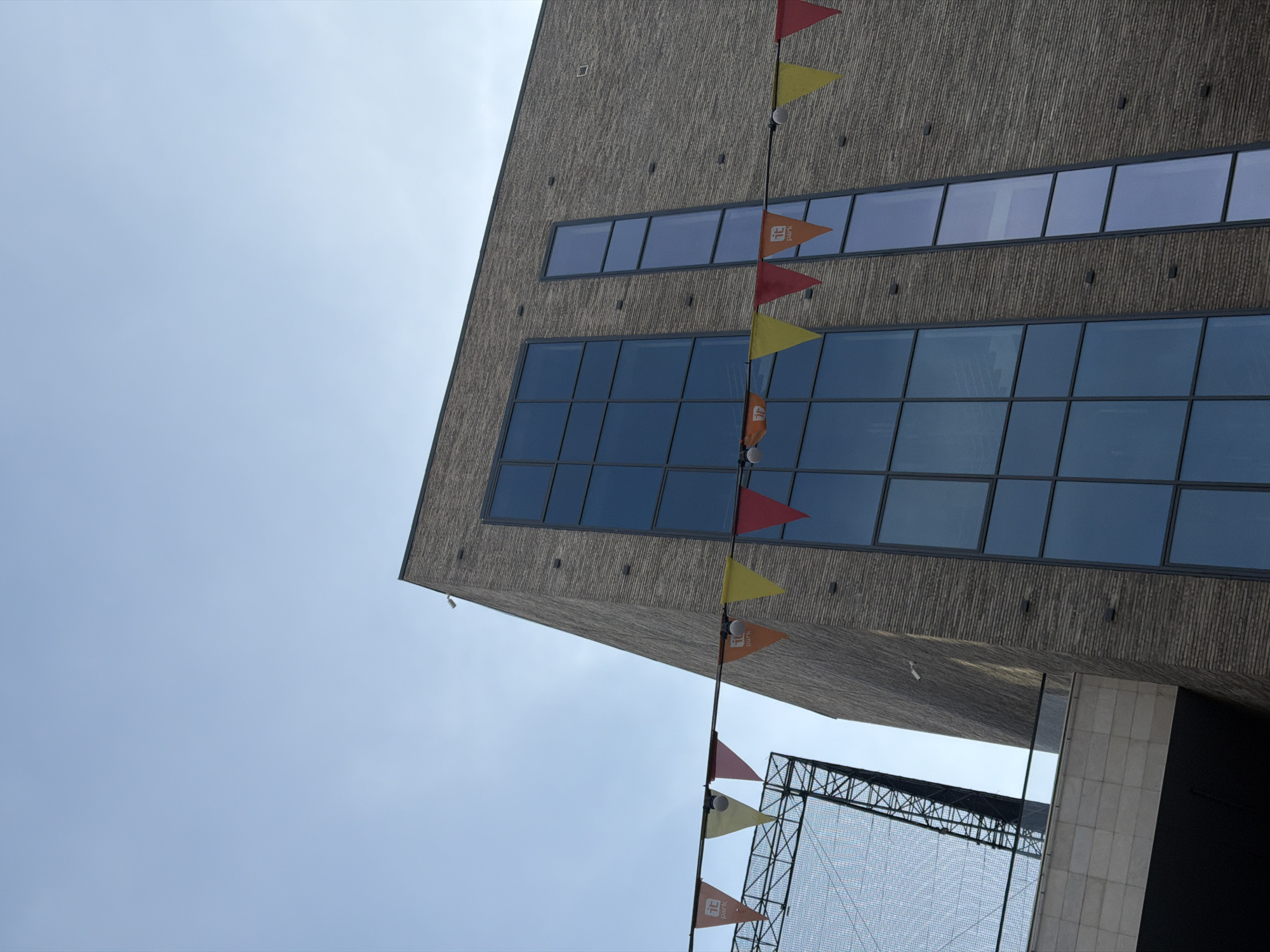 The image captures a portion of the IT Park building in Kazan, Russia, viewed from a low angle, looking upwards towards the sky. The building's facade features a rough, horizontally-patterned texture. Two rows of large windows, reflecting the sky, are visible on the building. A string of triangular flags in red, yellow, and orange with the IT Park logo hangs vertically down the facade. Below the main building, a metal structure with a mesh-like covering is partially visible, along with a corner of a light gray building. The sky is pale blue with slight cloudiness.