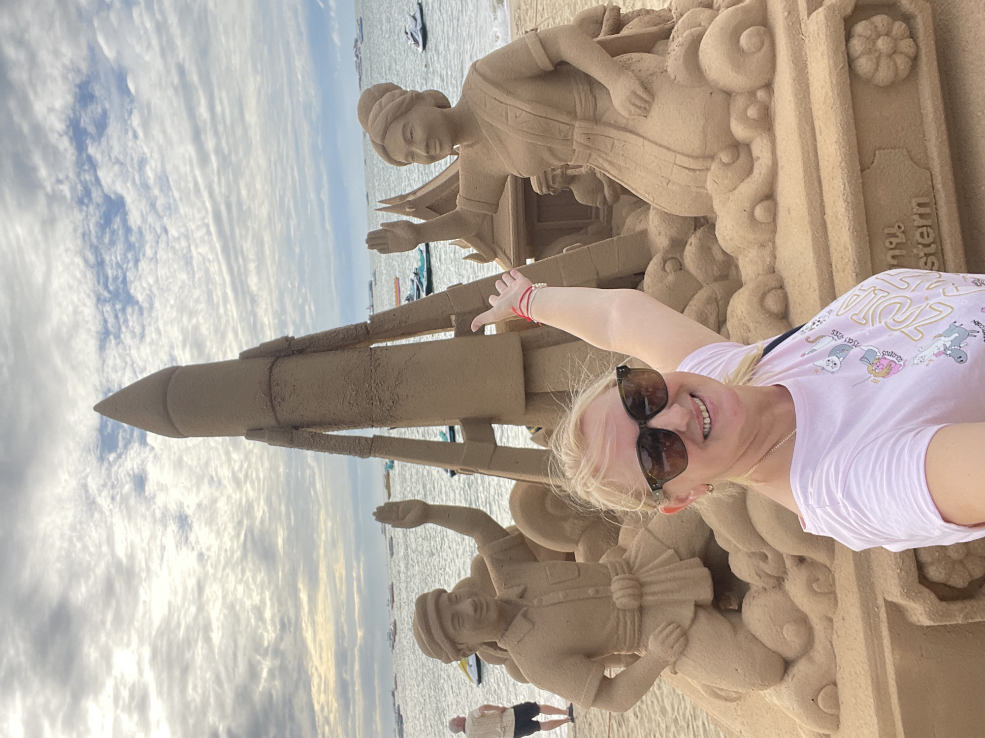 This is a portrait photograph taken by a woman standing on a beach in Pattaya City, Thailand, with a sand sculpture in the background. The woman is smiling at the camera, wearing sunglasses and a pink t-shirt. She is positioned in the foreground, partially obscuring the sand sculpture. The sculpture depicts a rocket on top of a tiered base, with two figures on either side. The figure on the left is a man kneeling with his arm raised, and the figure on the right is a woman kneeling with her arm raised. Behind the sculpture, the sea is visible with several boats and jet skis on the water. The sky is cloudy. The sand sculpture appears to be a significant attraction, possibly for tourists. The overall scene suggests a recreational setting at a beach resort.