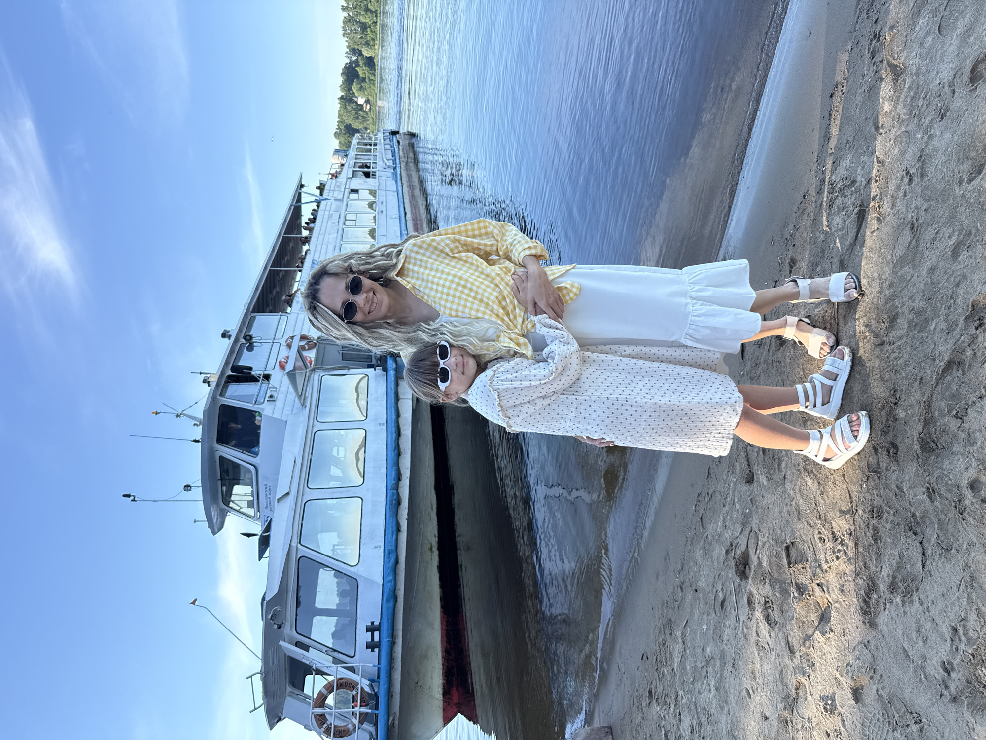 The image depicts two females, an adult and a child, standing on a sandy riverbank in Dumino, Russia. The adult female, with long blonde dreadlocks and dark oval sunglasses, wears a yellow and white checkered shirt over a white top, a white long skirt with a ruffled hem, and white flat sandals. She stands with her arms around the child. The child, with shoulder-length brown hair and white rectangular sunglasses, wears a white long-sleeved dress with small dark patterns and white platform sandals. She stands with her arms around the adult. Both individuals are smiling and looking towards the viewer.
Behind them, a large white passenger boat is docked in the calm, dark blue water. The boat features a blue stripe along its waterline and a red hull below it. Multiple windows are visible along its side, and an upper deck with railings shows several passengers. The inscription "Ярославский речной порт" (Yaroslavl River Port) is clearly visible in Cyrillic on the boat's forward superstructure. A white life buoy with partially visible Cyrillic text is attached to the boat's bow. The sky above is clear and bright blue, with minimal cloud cover. On the distant riverbank, a line of green trees and faint structures are visible. The foreground consists of light brown sand with numerous footprints. The scene is illuminated by bright, natural daylight, casting soft shadows on the sand.