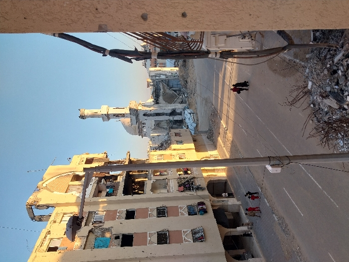 The image, captured from an elevated viewpoint, depicts a street in Khan Yunis, Palestinian Territory, showing an urban landscape marked by significant destruction. The scene is illuminated by the low sun, suggesting either early morning or late afternoon, casting long shadows across the road and buildings under a clear blue sky.
On the left side of the frame, a multi-story building stands severely damaged. Large sections of its facade are missing, exposing internal structures, broken windows, and rubble. Despite the extensive damage, blue tarps or fabrics are visible covering some openings, and clothes are seen hanging from windows or balconies, indicating a persistent human presence or attempts at salvage.
A paved road runs horizontally through the middle ground, bearing faint white lane markings. Several individuals are walking on the road. In the upper-middle section, two people in dark clothing walk towards the left. Closer to the foreground, a group of about three to four people, some dressed in red, also traverses the street. The road appears relatively clear, suggesting it remains in use. A utility pole or street light fixture extends from the left, over the road.
To the right of the road, a vast expanse of debris, including twisted rebar, concrete fragments, and rubble, signifies the complete demolition of structures that once stood there.
In the background, a mosque with a distinctive white dome and a tall, slender minaret is visible. It appears largely intact compared to the surrounding destruction.
No discernible text is visible within the image. The photograph conveys a profound sense of devastation and the continuation of daily life amidst the ruins of a conflict-affected area.
