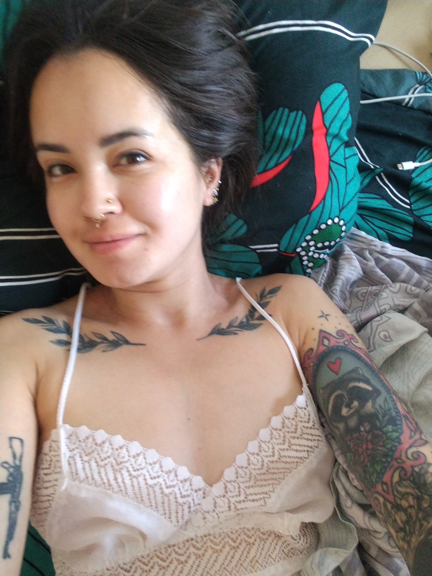 A woman with dark hair is lying down. She is wearing a white camisole with lace trim. She has tattoos on both arms and on her chest. Her left arm has a tattoo of a gun, and her right arm has a tattoo of a raccoon with a heart above it. She has two leaf-like tattoos on her upper chest/shoulder area. She has piercings in her nose and ears. She is lying on bedding with different patterns and colors. The location is not identifiable as Noviy, Russia from the image.