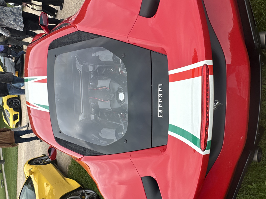 The image shows the rear end of a red Ferrari sports car, with a white and green racing stripe, parked in a grass lot. The car's engine is visible through a transparent cover. The car is parked near other cars, including a yellow sports car, and several people standing around. The location is near the Chateau de Chambord, a large French castle, located at 47.616 latitude, 1.518 longitude. The car is facing away from the camera. The camera is positioned to the right of the car, taking a rear angle shot. The Ferrari is the primary subject of the image, and it is in focus. The background is blurry, and the people are out of focus. There are no other details that can be gleaned from the image.