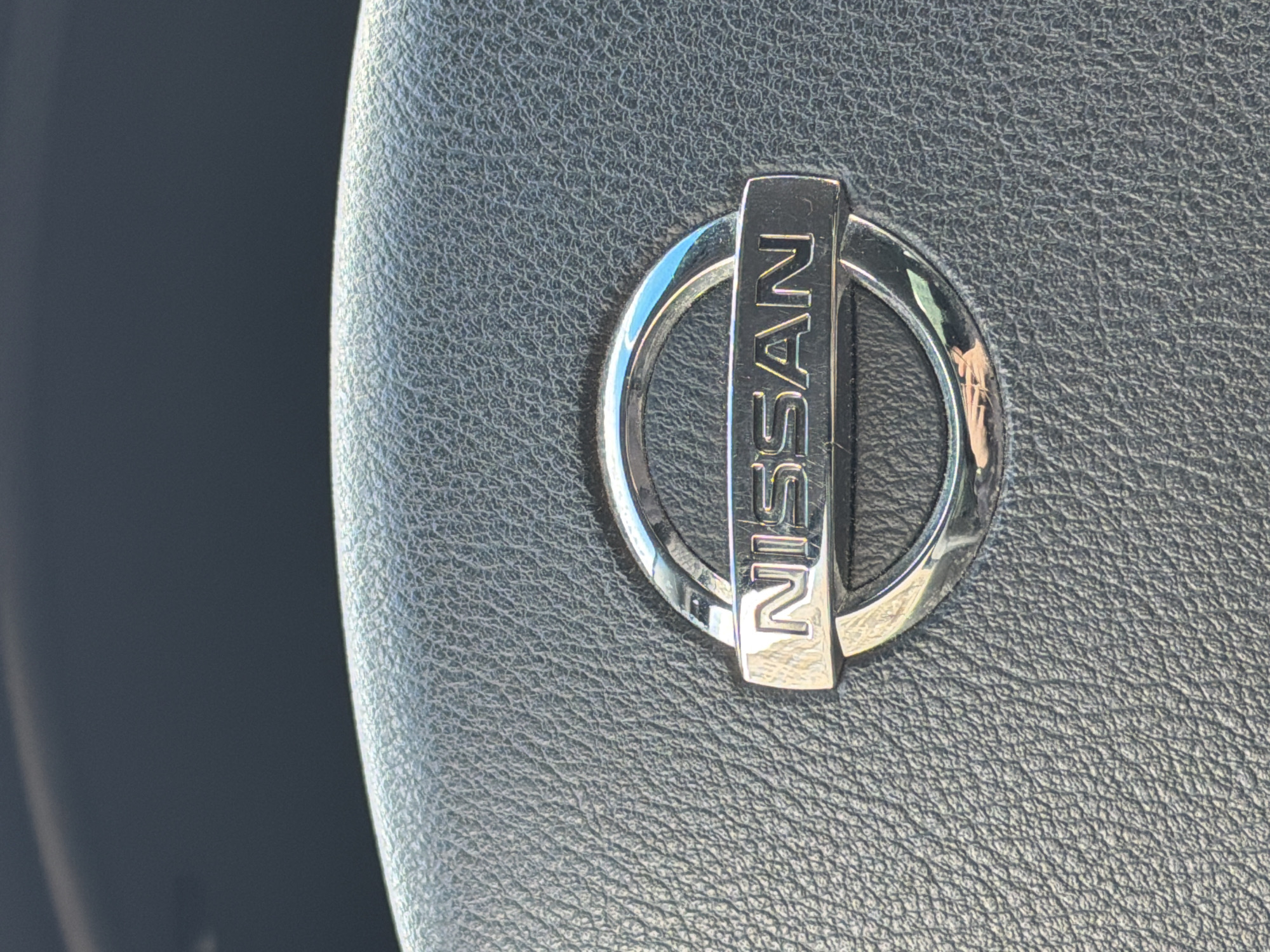 The image shows a close-up of a Nissan logo embedded in what appears to be a textured surface, likely the steering wheel or dashboard of a car. The logo is chrome and features the word "NISSAN" written vertically across a horizontal bar that intersects a circular ring. The surface surrounding the logo has a textured, leather-like appearance. The background is out of focus, suggesting a depth of field that emphasizes the logo. The image does not reveal any information about the city of Izhevsk, Russia.
