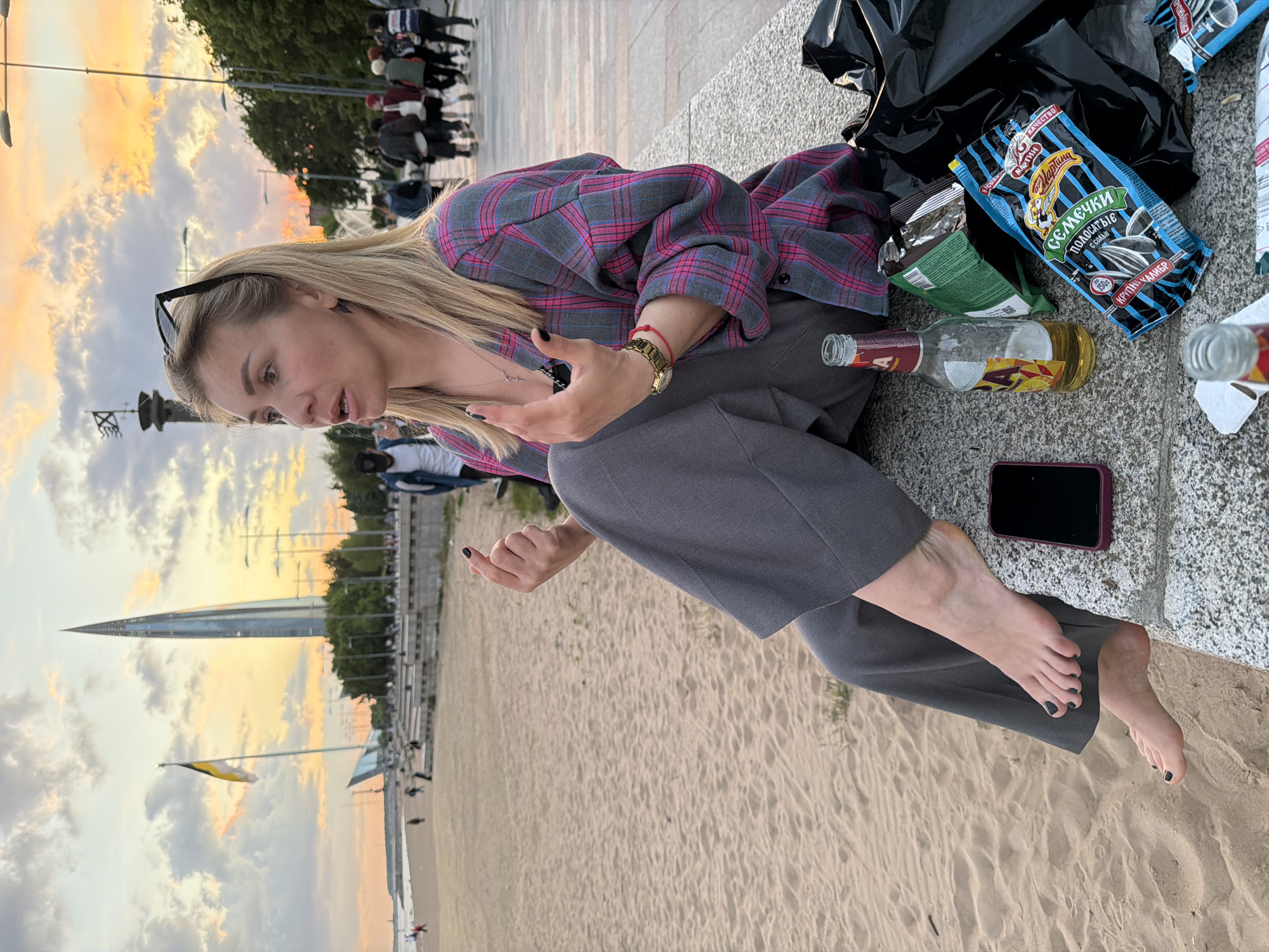 The image depicts a casual outdoor scene at sunset or dusk, focusing on a young woman sitting on a concrete embankment beside a sandy beach. The setting is identified as Saint Petersburg, Russia, due to the prominent Lakhta Center skyscraper in the background.
The main subject is a blonde woman, likely in her 20s or 30s, seen from a side profile. She is barefoot, wearing grey wide-leg trousers and a grey, pink, and blue plaid button-up shirt. Her hair is long and blonde, and sunglasses are perched on top of her head. She wears a gold watch, a red string bracelet, and a small cross necklace. Her fingernails and toenails are painted dark. Her mouth is slightly open, and her gaze is directed away from the camera, suggesting she is speaking or reacting to something.
Beside her on the concrete embankment are several objects indicating a relaxed gathering. These include a partially empty glass bottle with yellow liquid residue, a crumpled black plastic bag, and multiple snack bags. One bag of sunflower seeds is clearly visible, labeled "СЕМЕЧКИ ПОЛОСАТЫЕ ОТ МАРТИНА" (Striped Sunflower Seeds by Martina), with additional text "КРУПНЫЙ КАЛИБР" (Large Size) and "150г", along with "КАЧЕСТВО" (Quality). Another partially seen green bag appears to be a snack brand, possibly "СЕЛЯНОЧКА". A dark-colored smartphone with a purple case rests near her feet.
The time of day is evident from the warm, golden and orange hues of the sky, indicating late afternoon or early evening. The weather appears partly cloudy. In the distance, beyond the sandy beach, the distinctive, pointed silhouette of the Lakhta Center skyscraper dominates the skyline. A flag, yellow and white/light blue striped, flies from a pole further down the embankment. The sandy ground extends from the foreground, suggesting a beach or shoreline area, with faint figures of other people visible in the distance along the pathway and near the water. The scene conveys a sense of informal leisure by the water's edge.