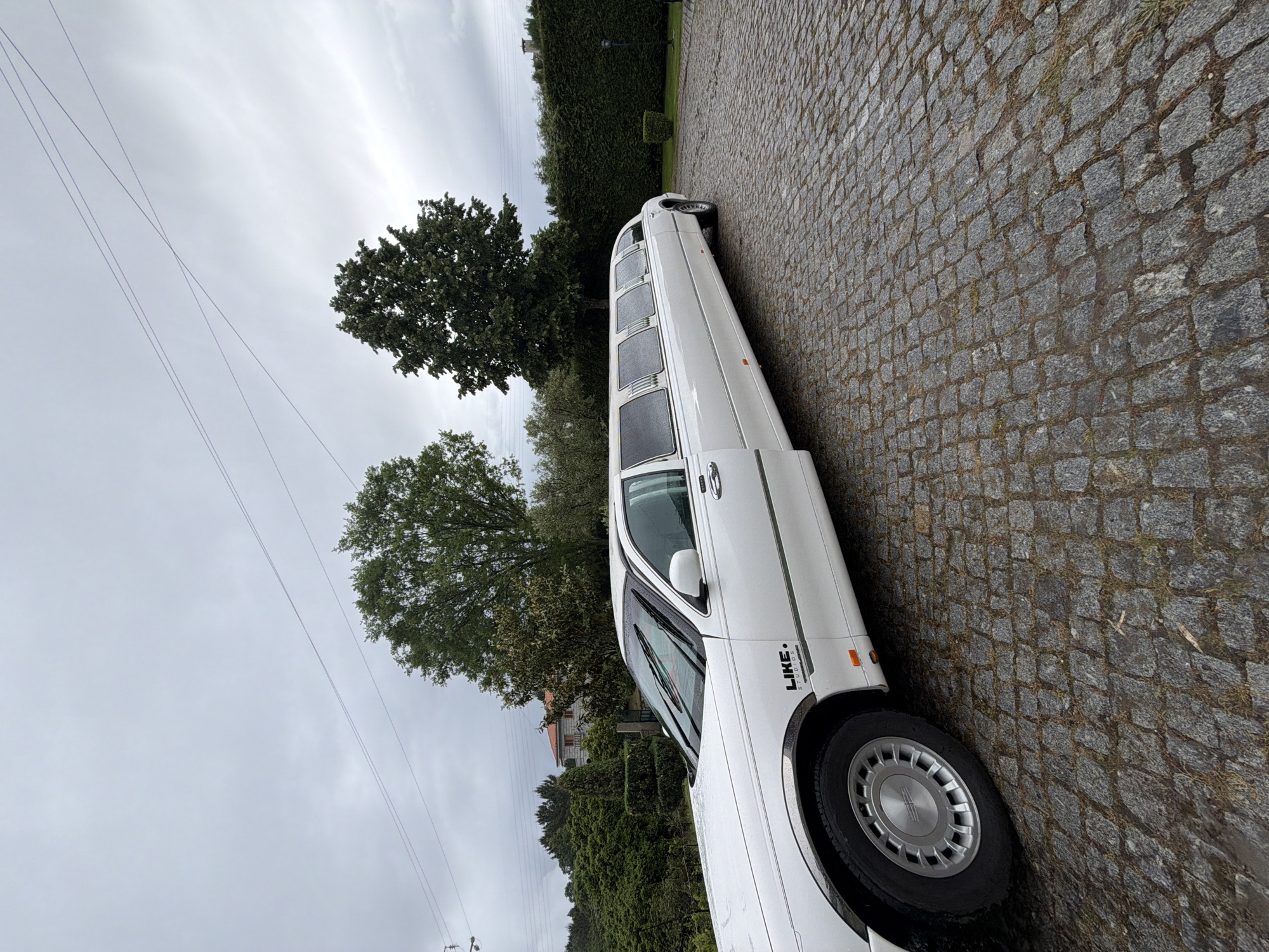 A white limousine is parked on a cobblestone road in Vila Real, Portugal. The location is suggested by the vegetation and architectural styles, which are typical of the region. The sky is overcast. A power line runs diagonally across the upper left portion of the frame. A few trees are visible. A portion of a building is barely visible behind the shrubbery. "LIKE. STUDIOS" is written on the side of the vehicle. The photo is taken at an angle.