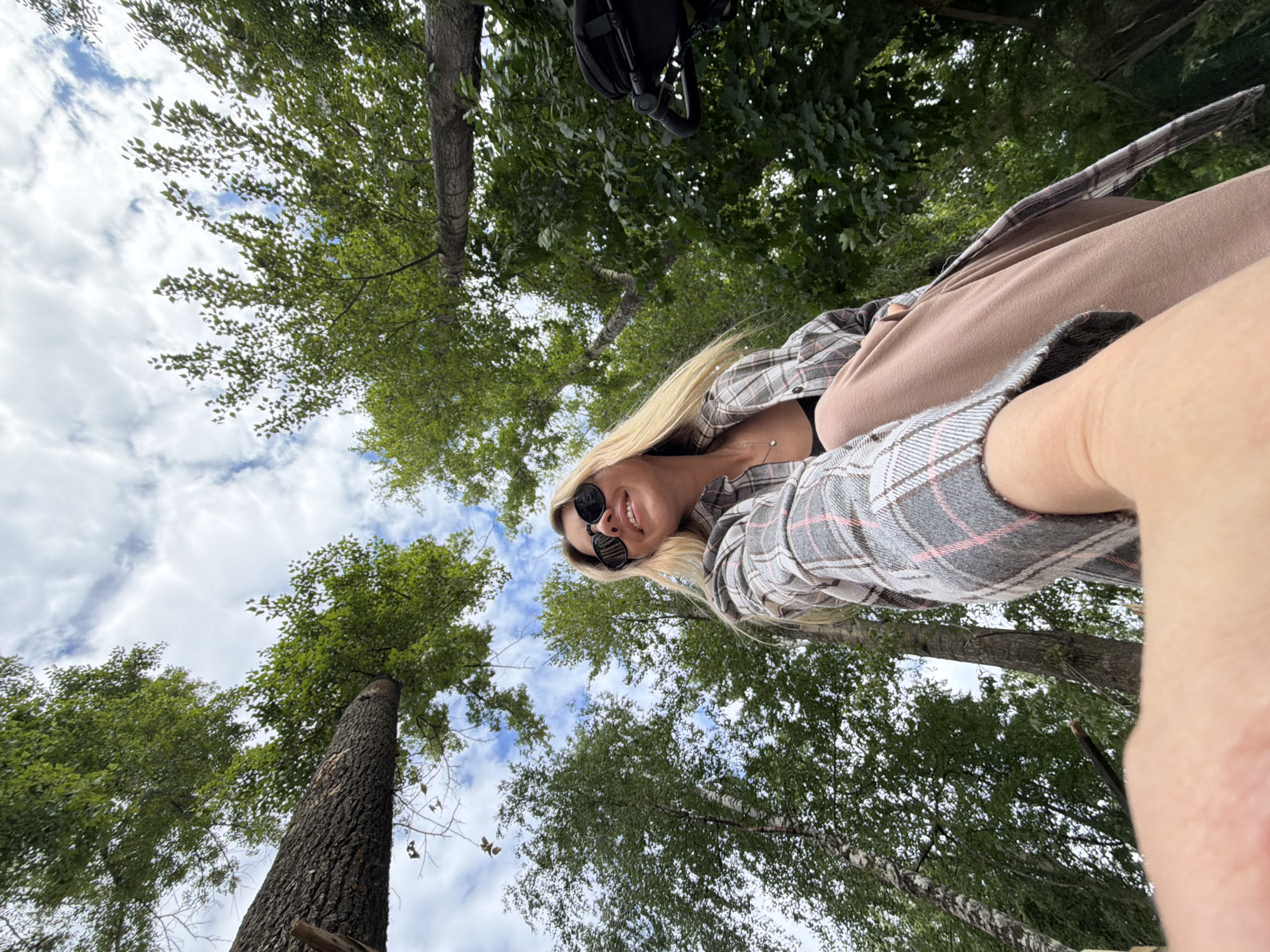 The image is a low-angle, wide-shot selfie taken outdoors in Chaikovsky, Russia, likely in a park or wooded area. A young woman with blonde hair and sunglasses is looking up and smiling. She is wearing a plaid shirt over a dark top and light-colored pants. Her right arm is extended, holding the camera. In the background, lush green trees with a partly cloudy sky are visible, suggesting it's daytime. A dark object, possibly a stroller or backpack, is partially visible on the right side of the frame. The overall setting is natural and casual.