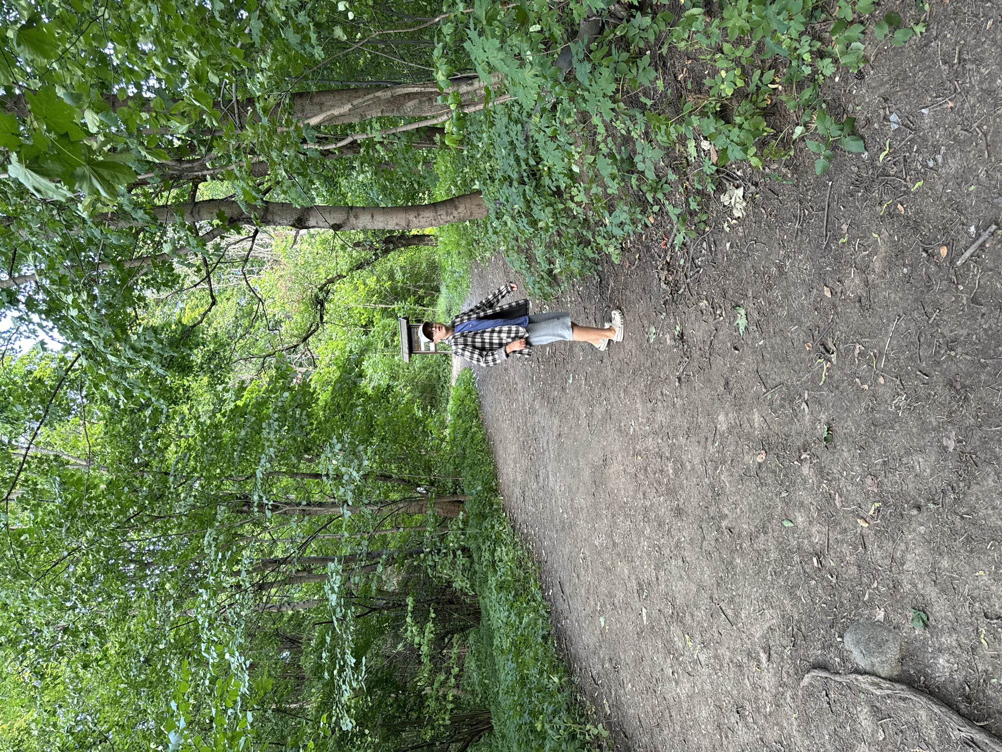 A young person wearing a plaid jacket, blue t-shirt, grey shorts, and white sandals stands on a dirt path in a wooded area. The path curves to the left and is surrounded by lush green trees and foliage. In the background, a wooden signpost with a white top is visible. The ground is covered in dirt, leaves, and small stones. The lighting suggests it is daytime.