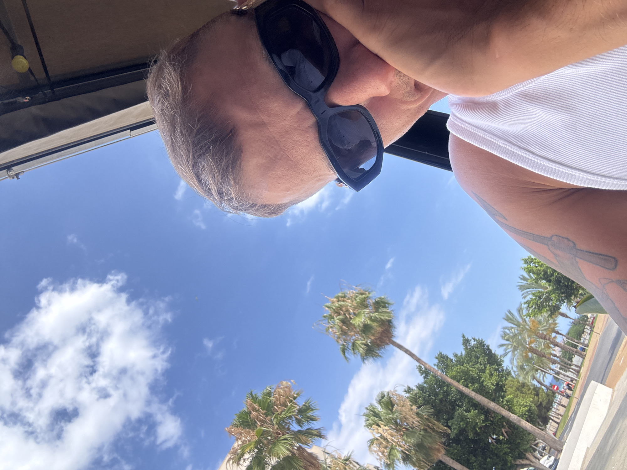 The image is a low-angle, slightly tilted, close-up shot of a person from the chest up, looking down at the camera. The person is wearing large, dark sunglasses and a white ribbed tank top. Part of their arm is visible on the right, with a tattoo of what appears to be a sword or dagger. The background is a bright blue sky with scattered white clouds. In the distance, several palm trees are visible, suggesting a warm, sunny location. The overall impression is a casual, summery scene, possibly taken outdoors in a warm climate like La Vila Joiosa, Spain. The lighting and bright sky indicate it is daytime, likely midday. No visible text is present in the image.