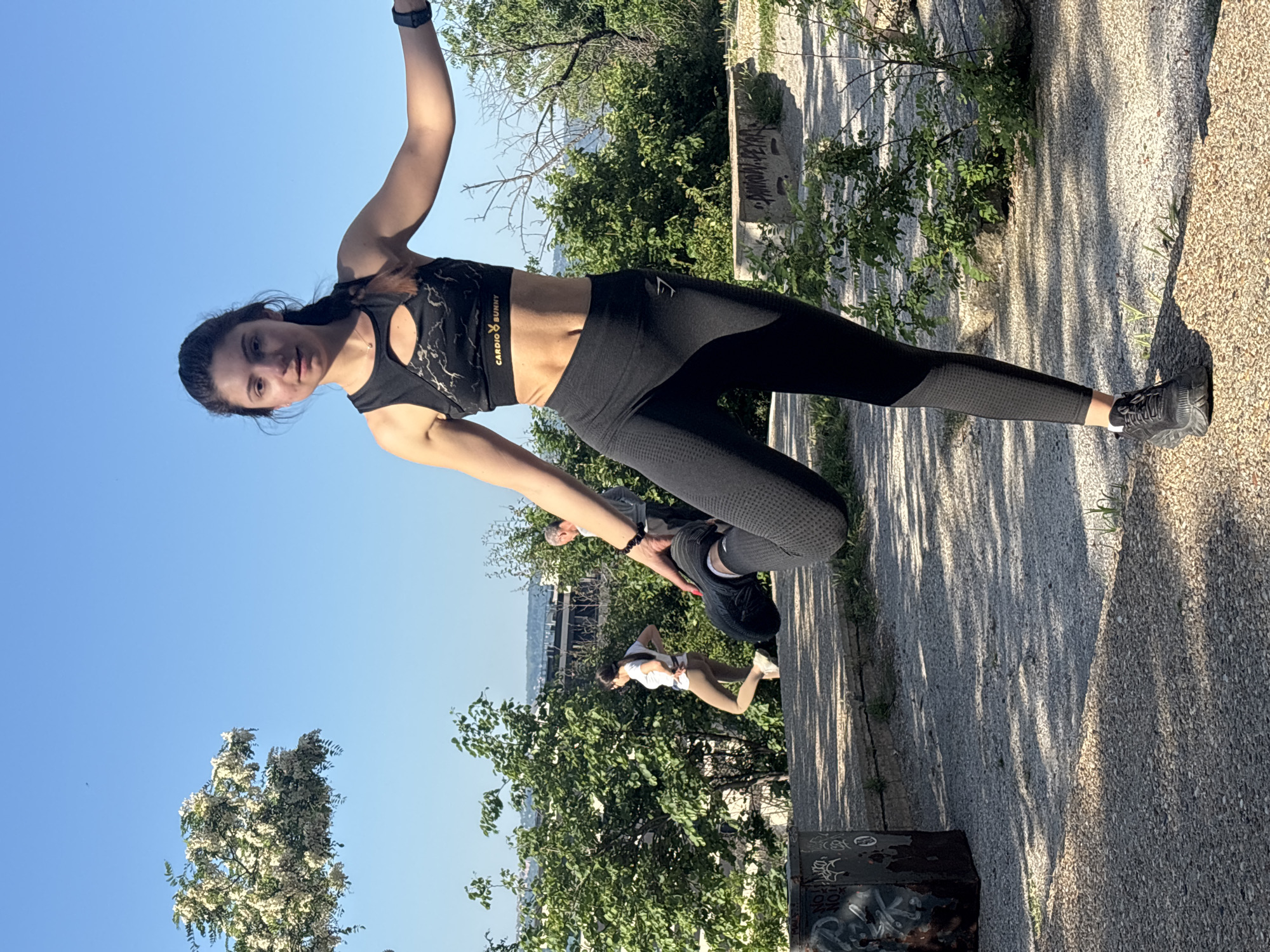 A woman in a black sports bra and leggings is standing on a gray sidewalk in what appears to be a park in Varna, Bulgaria. The park has lush green trees and shrubs, and there is a concrete structure with graffiti on it. A man and a woman are walking in the background. The sky is blue.