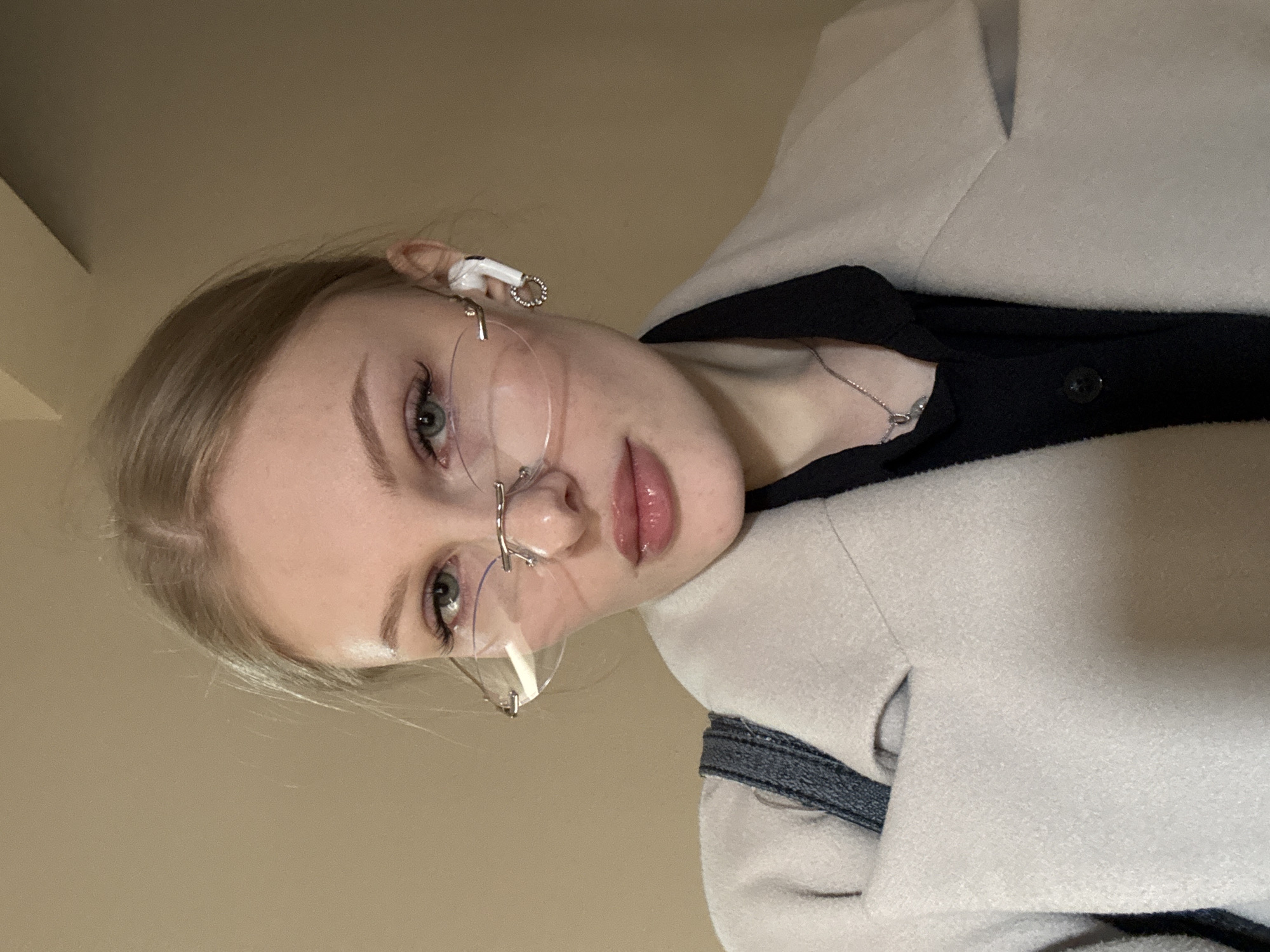 A young woman with blonde hair and glasses, wearing a white coat and a black shirt, is visible. She is likely indoors, given the lighting and background. A small earring is visible on the right ear. The location is unknown, and no connection to Perm, Russia is apparent.
