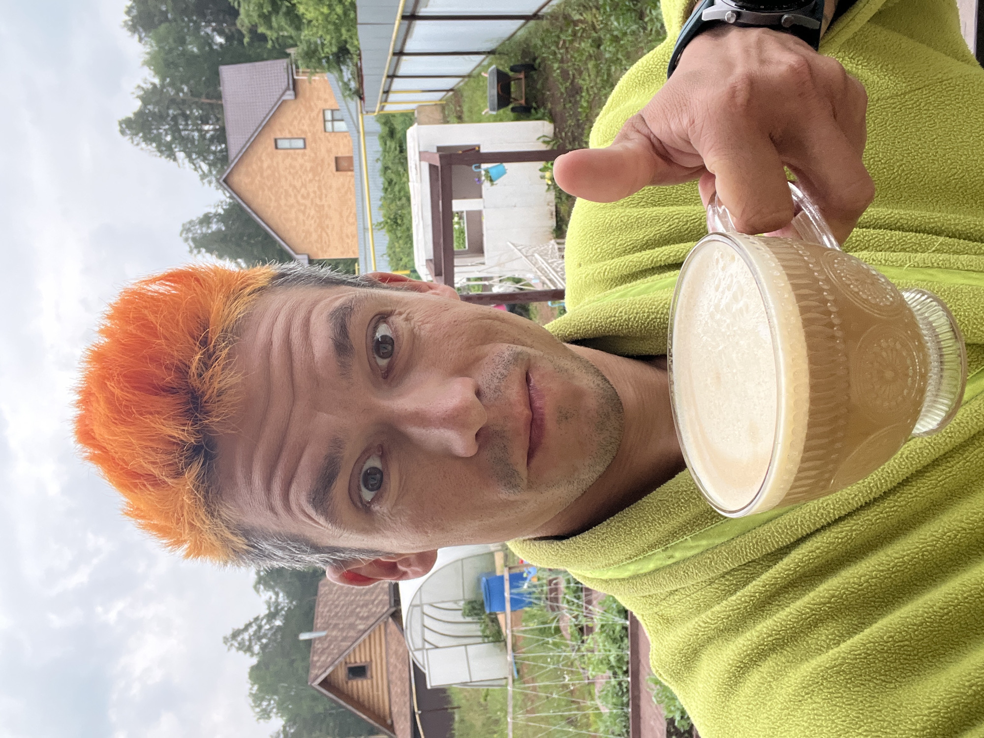 A man with bright orange spiky hair, wearing a lime green bathrobe, takes a selfie in an outdoor setting. He is holding a clear glass cup of coffee with foam in his right hand, while giving a thumbs-up with the same hand. A wristwatch is visible on his left wrist. In the background, there are houses, a greenhouse, a fence, and trees, suggesting a rural or suburban environment, possibly on the outskirts of Chaikovsky, Russia. The sky is overcast.