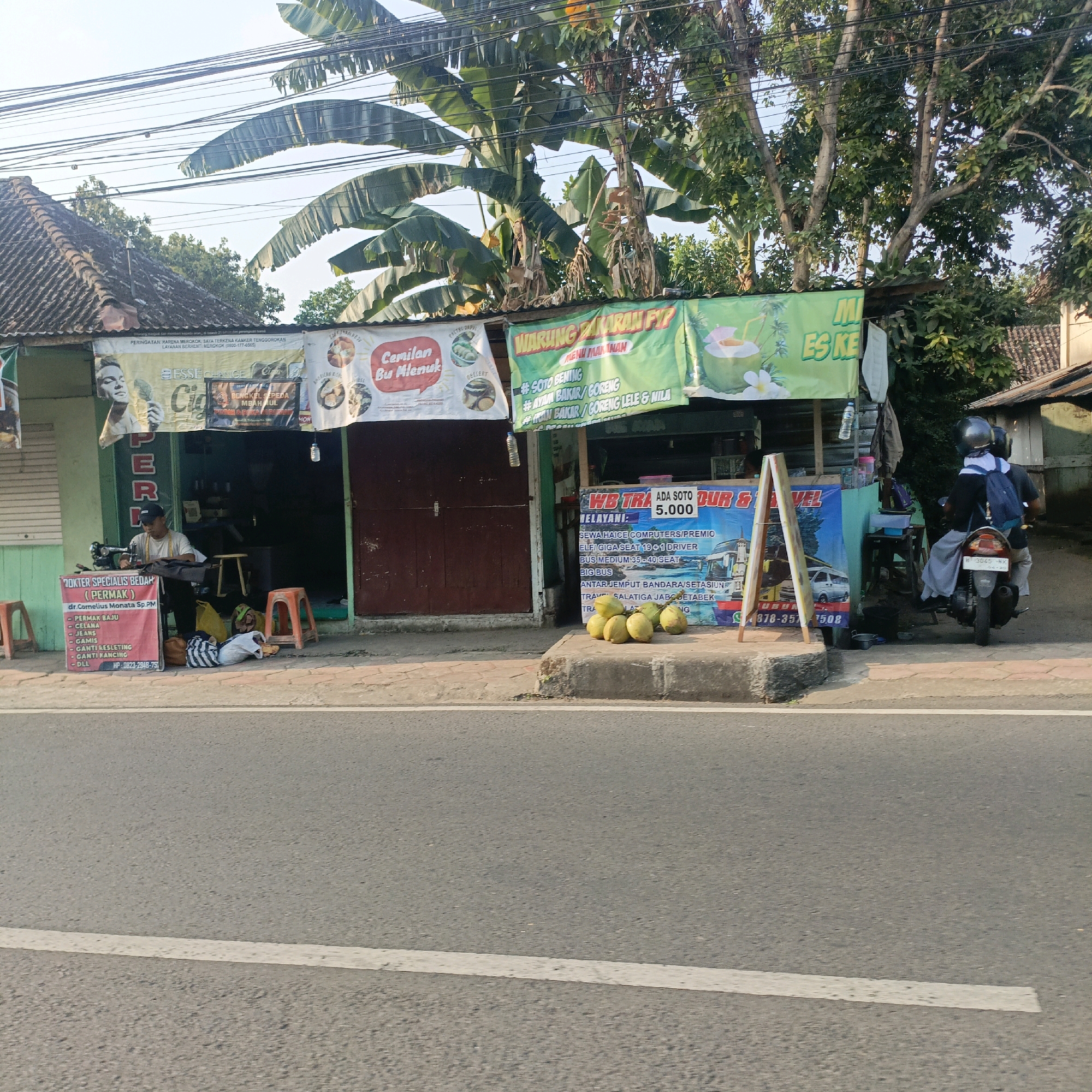 This image depicts a street scene in Salatiga, Indonesia, featuring a row of small businesses operating from open-fronted structures. The foreground shows a paved road with a white line marking. To the right, a sidewalk area is visible, with a person on a motorcycle wearing a helmet and a backpack, indicating they are a passenger. The businesses lining the sidewalk include a tailor shop, identified by a sign offering "DOKTER SPESIALIS BEDAH (PERMAK)" and a sewing machine operated by a man. Next to it, a food stall or warung advertises menu items like "SOTO BENING" and "AYAM BAKAR/GORENG". Another stall displays a sign for "WB TRA" and "TOUR & TRAVEL", also advertising "ADA SOTO 5.000". Several coconuts are displayed on the sidewalk in front of this stall. Above the stalls, banana trees and other foliage are visible, along with power lines crisscrossing the sky. The overall impression is a vibrant, bustling street scene characteristic of a commercial area in Salatiga.