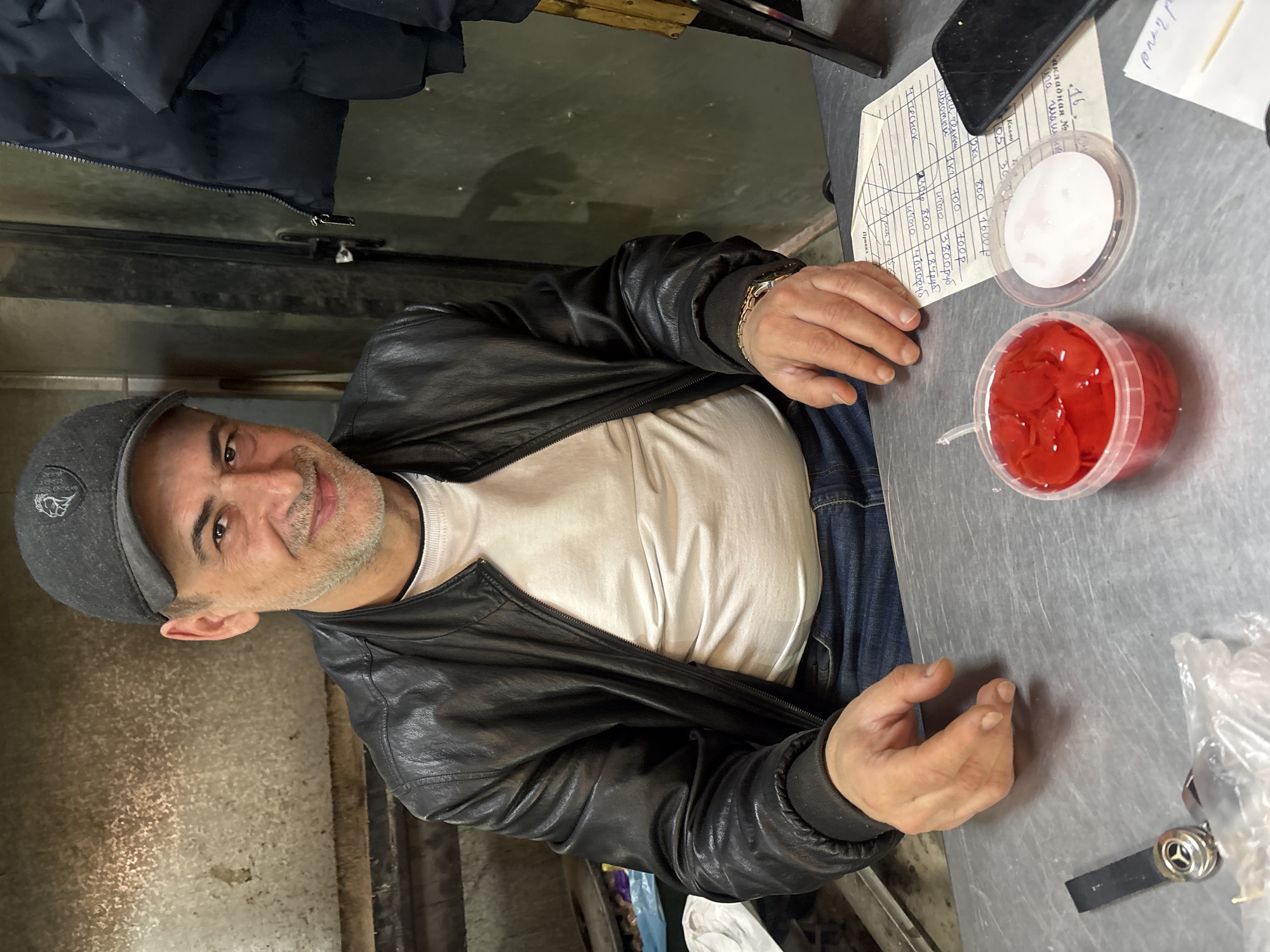 A middle-aged man with a gray cap and a black leather jacket sits at a metal counter. He has a light beard and is looking at the camera with a slight smile. On the counter, there's a sheet of paper with handwritten notes in Russian (likely an invoice), a black phone, and two plastic containers, one filled with a white substance and the other with red, cherry-like objects. The environment appears to be a business-like setting. There is no indication the location is in Krasnoyarsk, Russia.