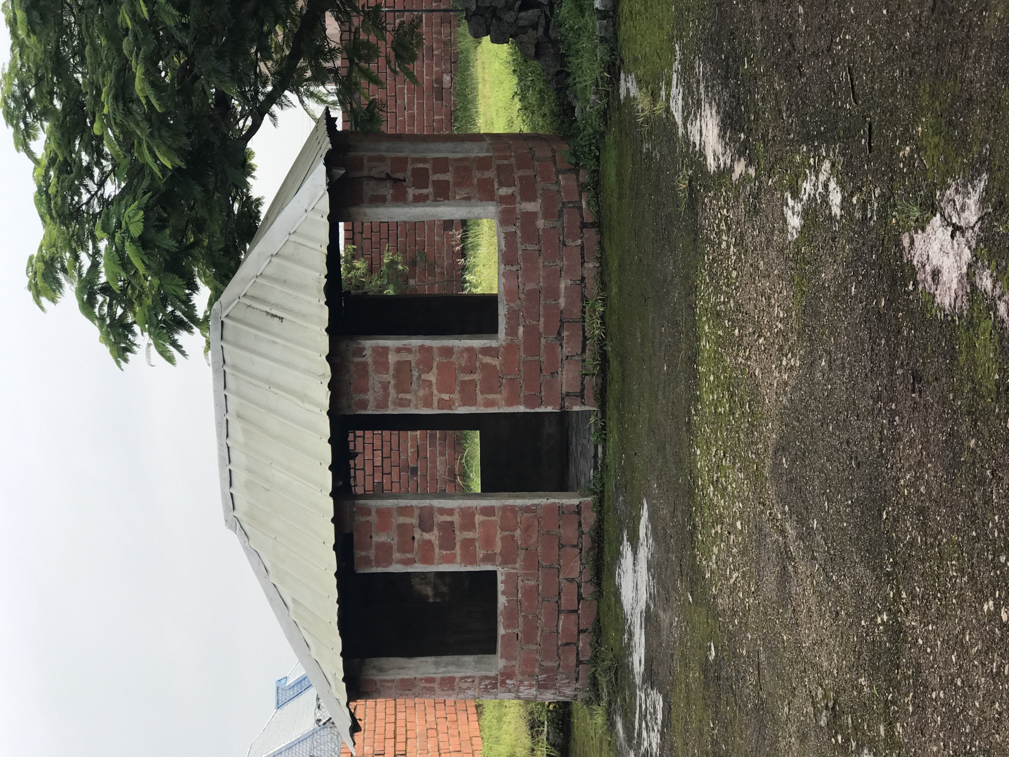 The image depicts an unfinished circular structure constructed from red bricks and concrete, with a corrugated metal roof. The structure has three openings, two of which appear to be windows and one is a large entrance. The surrounding area is grassy with patches of wet, muddy ground, and moss is visible on the ground and the lower parts of the structure. In the background, there is a brick wall and a large, leafy green tree. The sky is overcast and cloudy. The scene appears to be an outdoor location in Jos, Nigeria, possibly a construction site or an undeveloped area. There are no people or animals visible in the image, and no objects that suggest a specific activity or interaction.