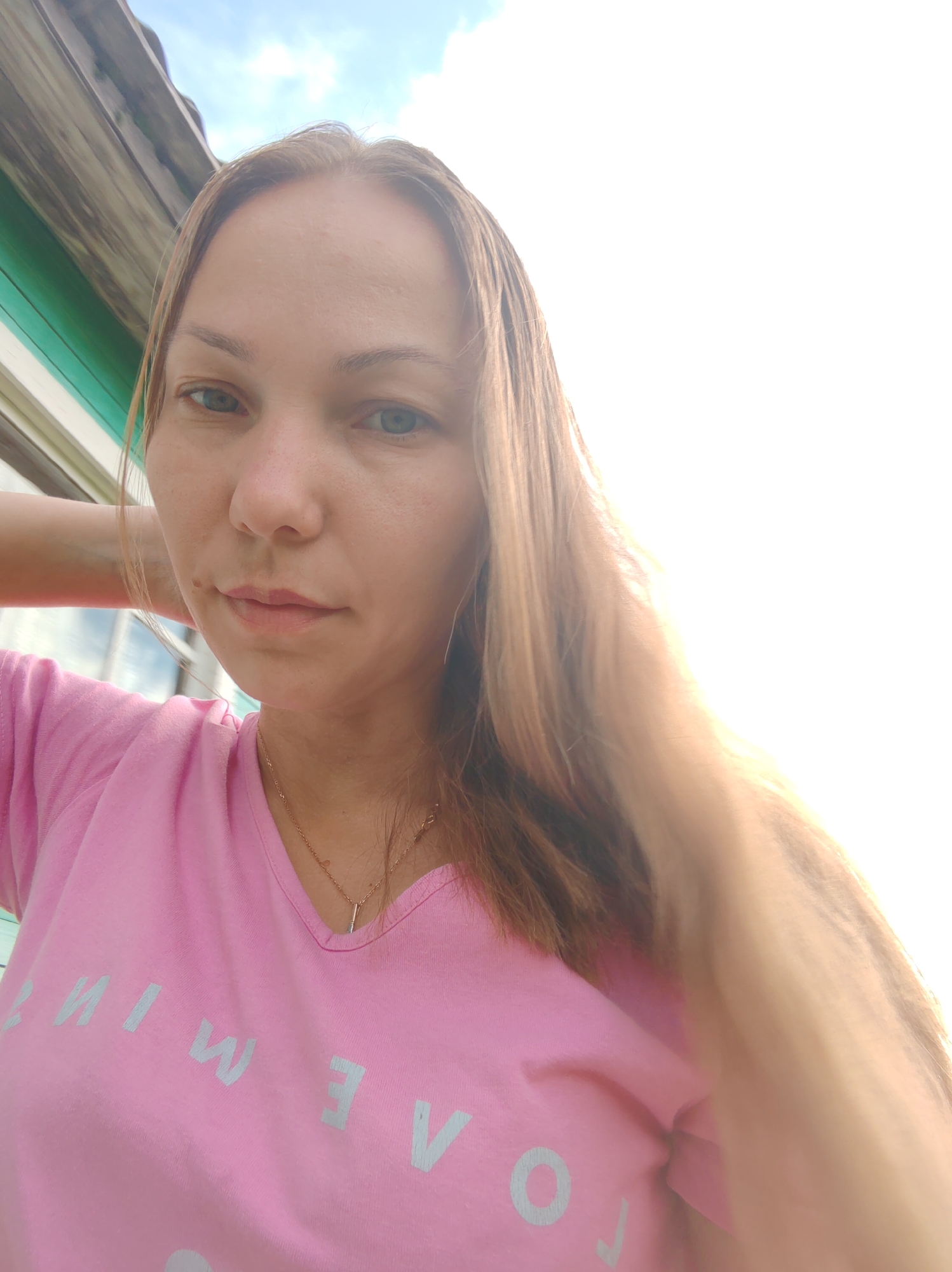 A woman is taking a selfie outdoors, with a bright sky behind her. She has light brown hair and is wearing a pink t-shirt. The t-shirt has some text on it, but it is partially obscured by her hair and the angle of the photo. She is holding her left hand up to her head, and her hair is being brushed to the right. In the background, there is a wooden structure with green trim, which could be part of a house or shed. The overall lighting suggests it is daytime, possibly late afternoon due to the warm, hazy light. The location is identified as Chukhlem, Russia.