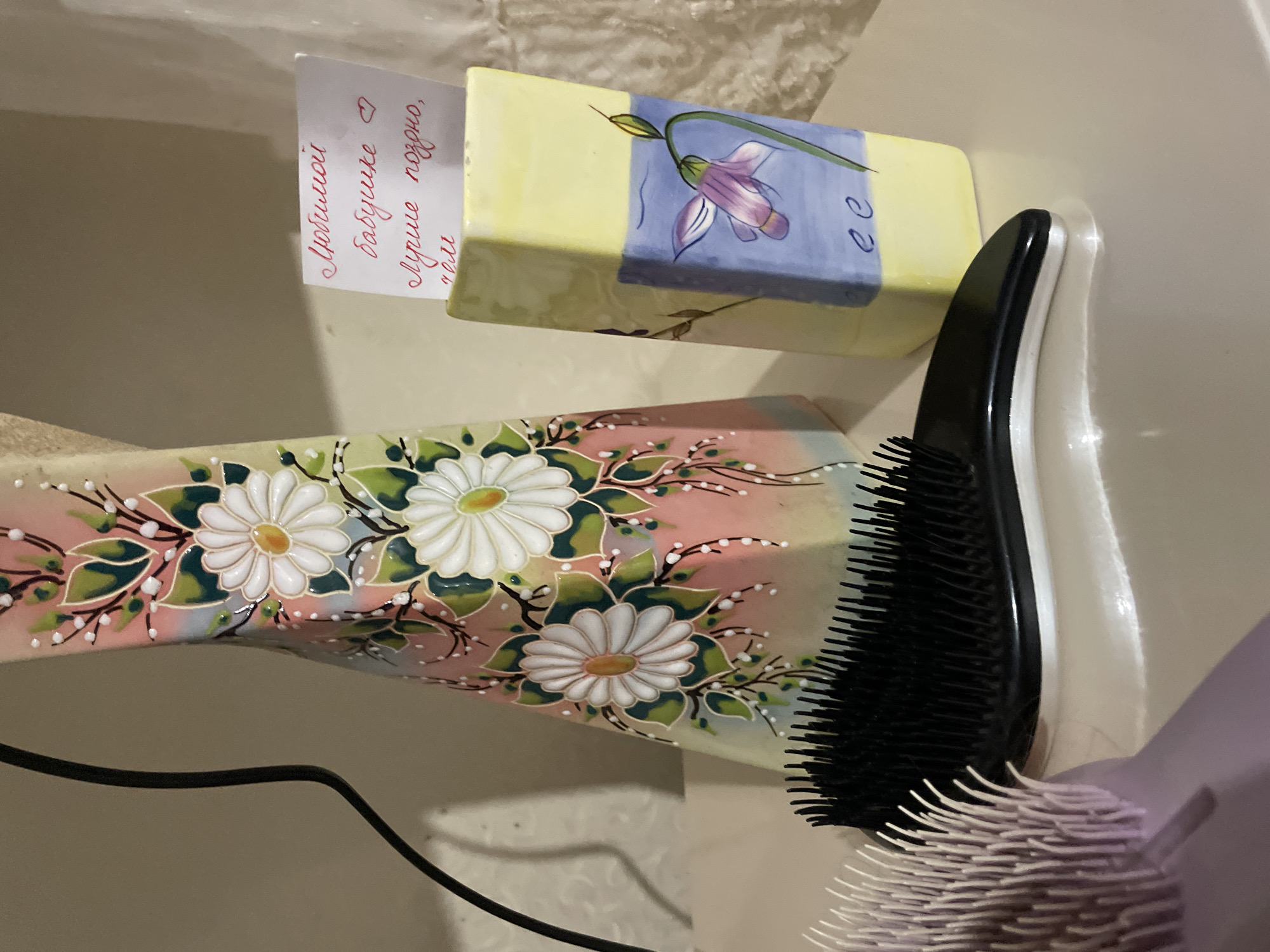 The image shows a collection of personal care items, possibly in a bathroom setting. A rectangular soap bar, decorated with a purple flower design, has a small white card inserted next to it. The card features red Cyrillic writing, possibly a personal message. Adjacent to the soap bar is a black hairbrush with a white handle. Below these items, there is a vase or container decorated with a pink and white daisy pattern. Part of a lilac-colored hairbrush is visible in the lower right corner of the image. A black power cable is visible in the lower left corner of the image. The backdrop is a smooth, light-colored surface, which could be part of a sink or counter. The location given is Olginka, Russia, though there is nothing in the image itself that conclusively indicates this location.