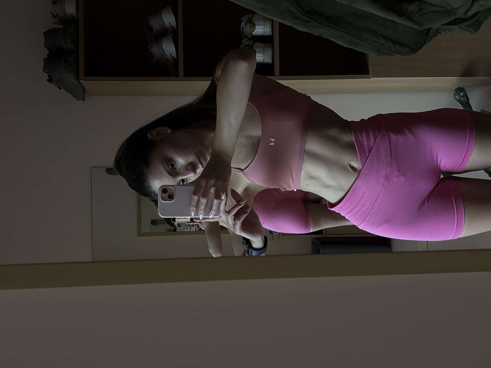 A woman with dark hair pulled back in a ponytail is taking a mirror selfie in what appears to be a bedroom or dressing area. She is wearing a pink sports bra and matching pink high-waisted shorts. Her midsection is visible, showing well-defined abdominal muscles. In the background, there is a tall mirror, a shelf unit containing several pairs of shoes, and a closet with a green garment hanging. The room is sparsely furnished and appears to be well-lit, possibly by natural light from a window. The location is not specified in the image.
