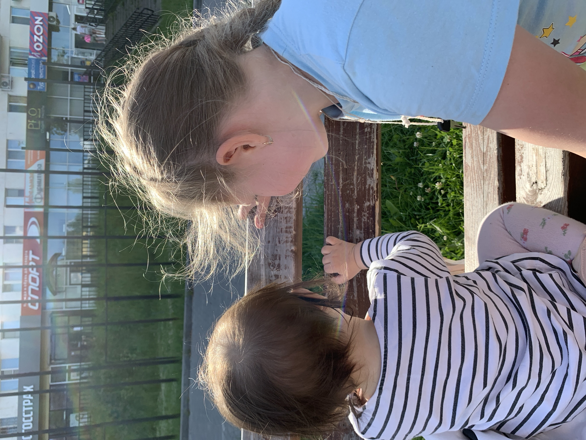 The image captures two young girls sitting on a wooden bench outdoors, facing away from the camera and towards a row of businesses in the city of Chaikovsky, Russia. The girl on the left has short, dark hair and wears a black and white striped long-sleeved shirt and light-colored leggings with a floral pattern. The girl on the right has long, blonde hair braided into a ponytail and wears a light blue t-shirt. Her right hand is visible resting on the bench. In the background, a fence separates the girls from a grassy area, and behind that, storefronts are visible with Russian signage. The sunlight casts a warm glow on the scene, and a slight rainbow effect is visible on the girl on the right's face. The overall environment suggests a casual, sunny day.