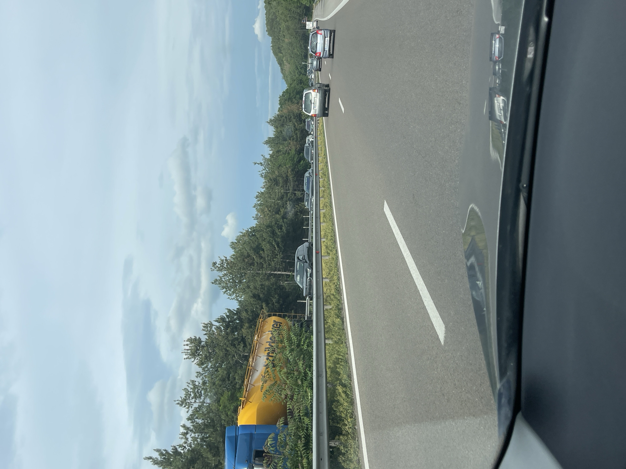 The image shows a highway with several cars driving, indicating traffic. On the left side of the highway, a large yellow and blue truck trailer is tipped on its side. The truck appears to be an industrial vehicle, possibly a cement or bulk material transporter, with the brand name "STRACKER" visible on its side. The trailer is partially obscured by foliage and a metal guardrail. The environment is characterized by a wooded area on the left side of the road, with a cloudy sky overhead. The road itself is paved and marked with white lines. The scene suggests a traffic incident, likely a rollover, has occurred. The location is identified as Gorno Varshilo, Bulgaria.