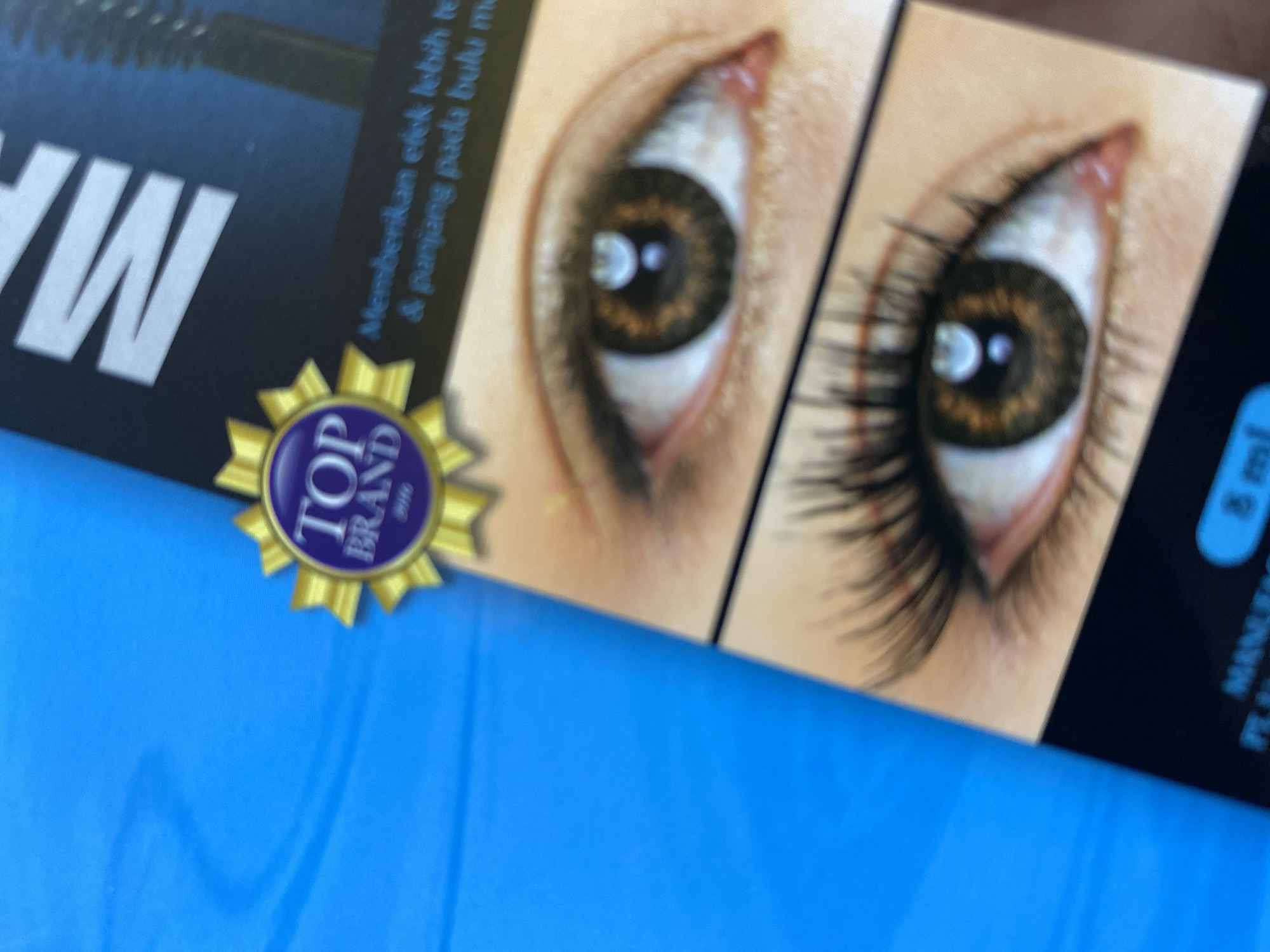 The image displays a close-up of a product box for mascara, featuring a "Top Brand" award and a visual comparison of before and after application on an eye. The product name "MASCARA" is partially visible in large white lettering at the top. The "Top Brand" award is a golden starburst with a purple center that reads "TOP BRAND" and "2016" below it. The product packaging also includes text in Indonesian, stating "Memberikan efek lebih tebal & panjang pada bulu mata" which translates to "Provides a thicker and longer effect on eyelashes." The image is angled and appears to be a product advertisement or packaging, possibly displayed on a blue surface. There is no identifiable location in Ta', Indonesia.
