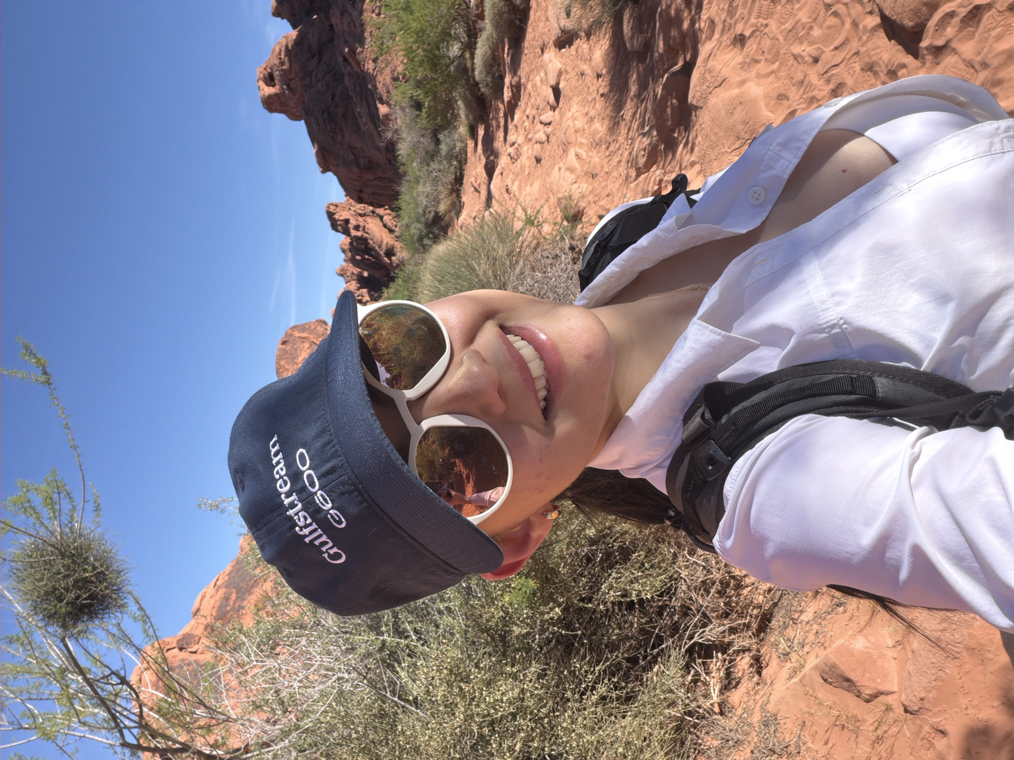 A medium-shot, slightly rotated selfie captures a woman in the landscape of Moapa Valley, United States. She wears a navy baseball cap, white-framed sunglasses, and a partially unbuttoned white shirt over a white top. A black backpack strap is visible on her right shoulder. The background consists of arid terrain with red rock formations, desert vegetation, and a clear blue sky. The woman is smiling, revealing her teeth. The reflection in her sunglasses shows someone in a similar attire.