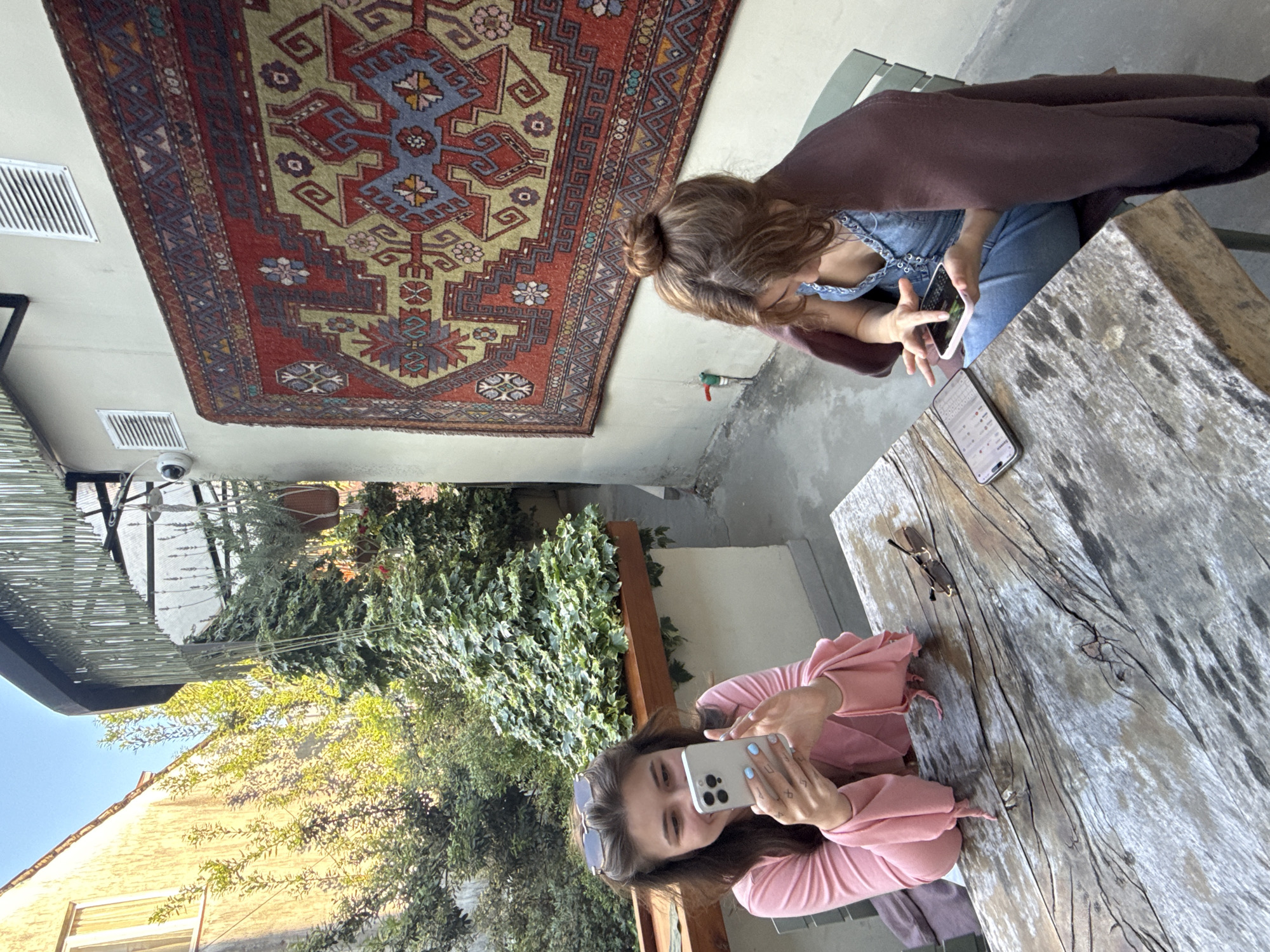 Two women are seated at a weathered wooden table in an outdoor patio or courtyard setting. One woman is wearing a denim top and has a brown shawl draped over her shoulders. She is holding a phone and appears to be interacting with it. The other woman is wearing a pink top and is also holding a phone, seemingly taking a picture or video. Sunglasses rest on the table.
Above the women, a large, decorative rug hangs on the wall. The patio is partially shaded by an awning and surrounded by lush greenery and building facades. The location is Batumi, Georgia.
