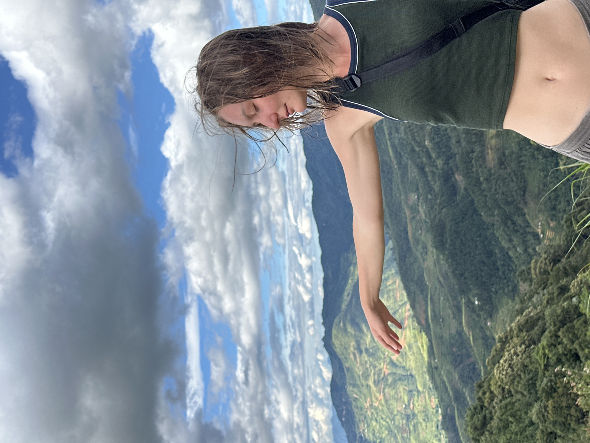 An outdoor vertical image depicts a young adult female against a mountainous backdrop in Hanh Phuc, Vietnam. The subject, positioned with her body angled slightly towards the right of the frame, has light brown hair that appears wind-blown, with her eyes closed and a calm expression. She wears a dark green ribbed tank top with white piping around the neckline and armholes, exposing her midriff and navel. Grey lower garments are partially visible. A black strap crosses over her right shoulder. Her left arm is extended horizontally to the left, palm down, with fingers slightly curved. The background features extensive green mountain ranges, receding into the distance under a sky populated by a mix of white cumulus and darker grey clouds, interspersed with clear blue patches. Evidence of terraced cultivation and sparse structures can be observed on some distant slopes. Dense green foliage occupies the lower left foreground.