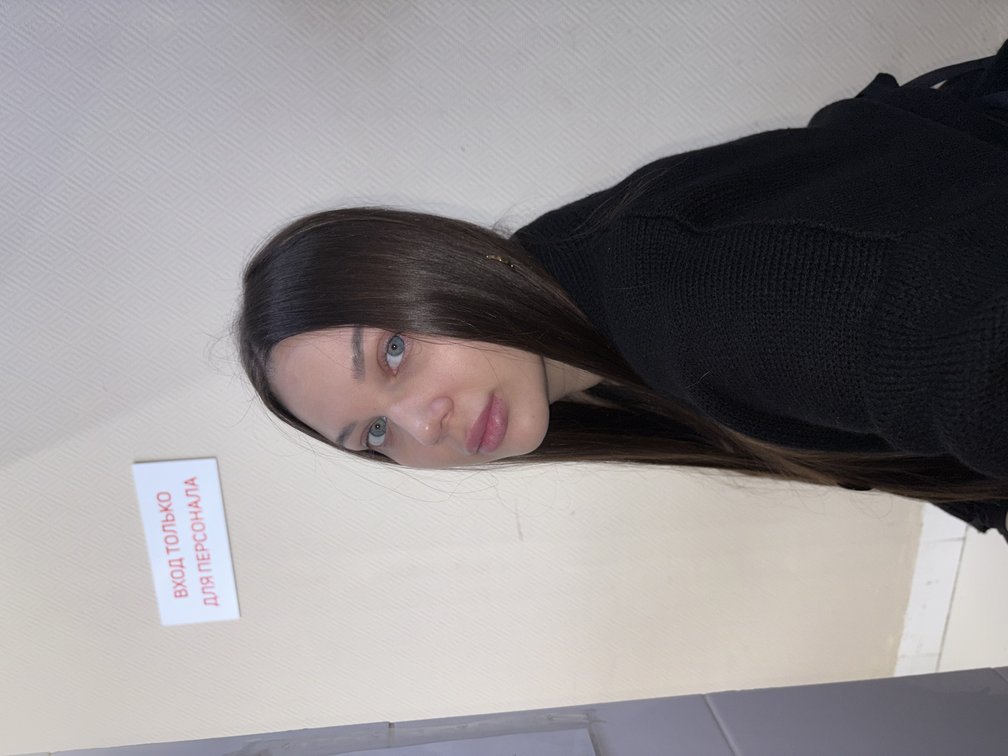 A woman with long brown hair and blue eyes is looking at the camera. She is wearing a black sweater. A sign with Russian text reading "Вход только для персонала" (Entrance for staff only) is visible on the wall. The location is likely the city of Chaikovsky, Russia. The woman appears to be indoors.