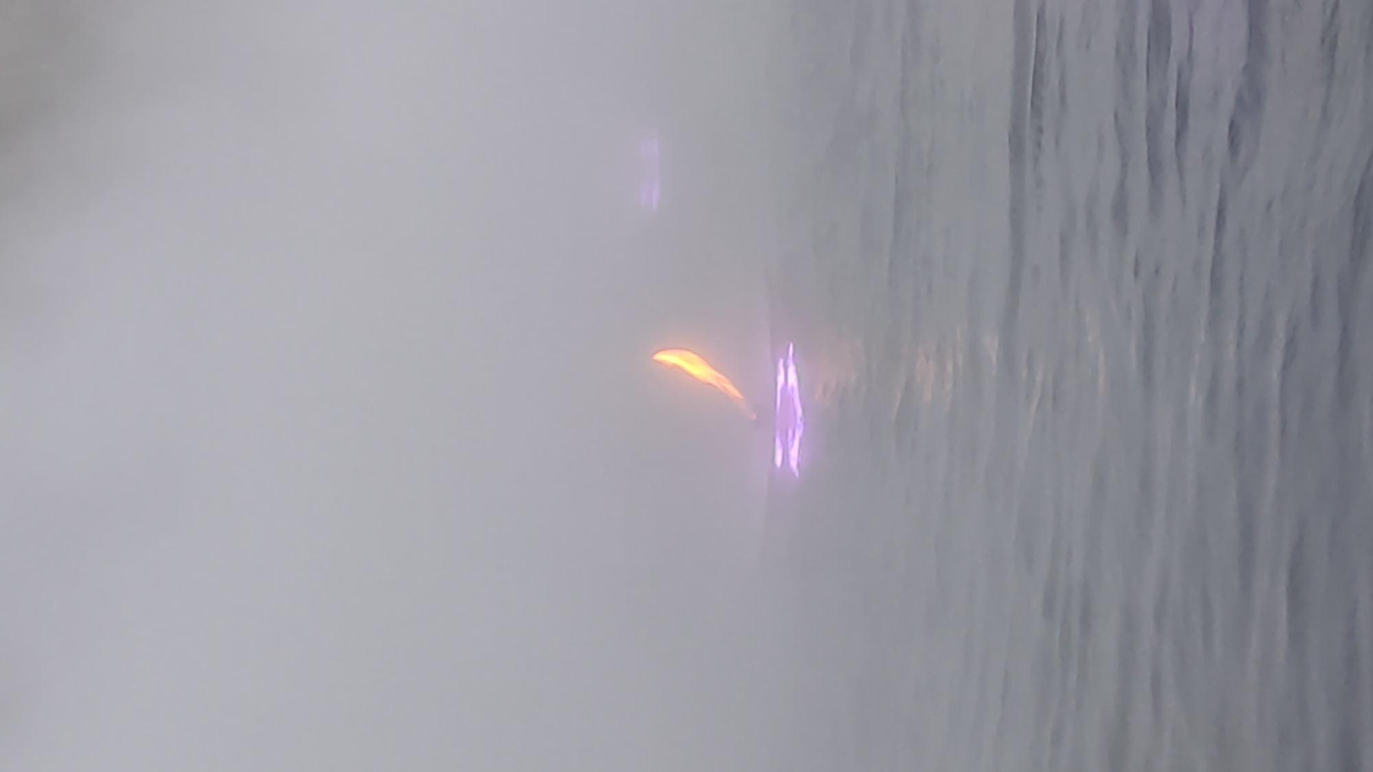 The image captures an atmospheric indoor scene dominated by mist and artificial light. A dense, grayish haze, resembling fog or steam, occupies the left and central parts of the frame, partially obscuring the background. Through this mist, two distinct light sources are visible. On the left, an elongated, curved orange-yellow light glows, reminiscent of a stylized flame or a narrow light fixture. To its right, a vibrant, abstract purple light appears, featuring an irregular, somewhat vertical shape, possibly a reflection or another decorative light. The right side of the image reveals a textured surface with vertical patterns, suggesting flowing water on glass or a similar water curtain feature. No people, discernible objects beyond the lights, or visible text are present. The time of day cannot be determined, but the artificial illumination implies an indoor setting, likely in the evening or night. The overall impression is one of a modern, possibly decorative, environment.