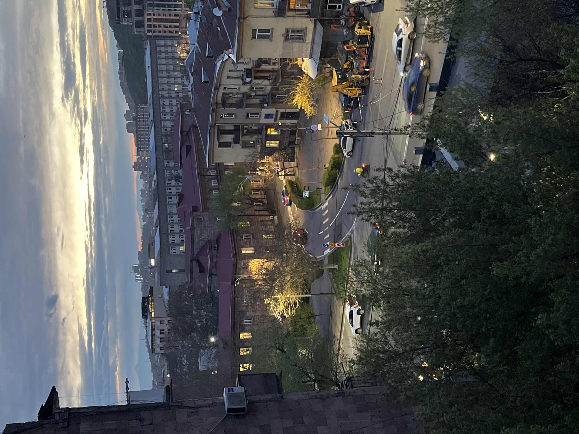 An elevated view of a street scene in Yerevan, Armenia, at dusk. Buildings of various heights and architectural styles line the street, illuminated by streetlights and lights from within the buildings. The sky transitions from a light blue to orange and gray. Traffic includes cars traveling along the street, with a construction site visible near the center of the frame. Trees are present in the foreground.
