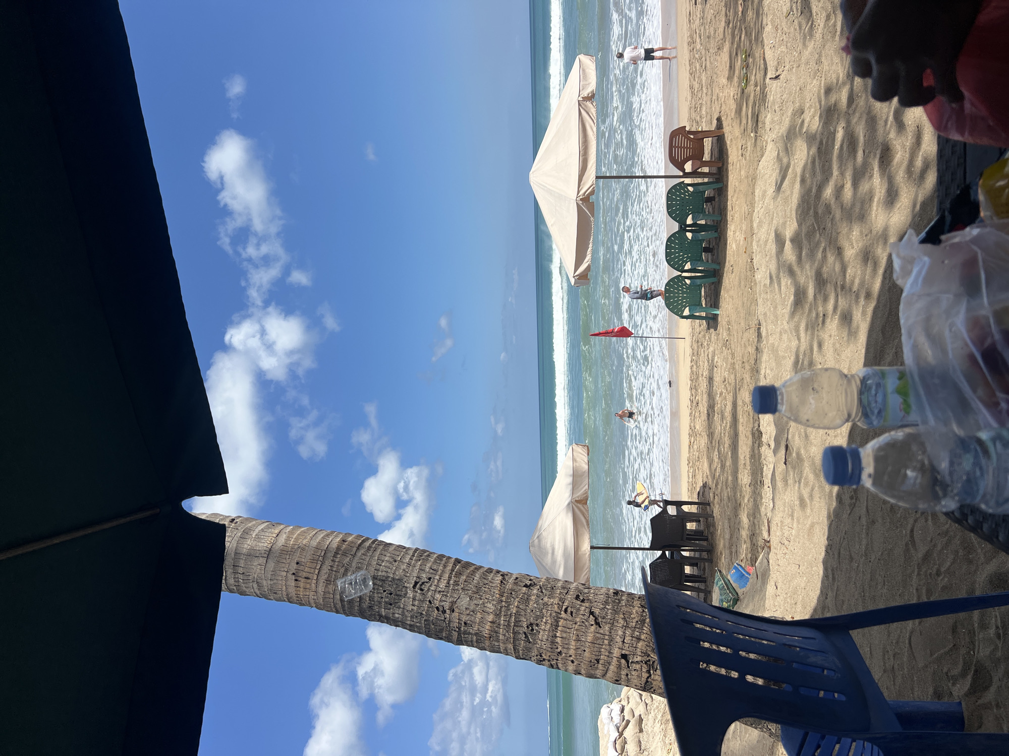 A beach scene in Kuta, Indonesia, features a sandy shore with people and beach chairs under umbrellas. A large palm tree stands to the left, with a plastic bottle tied to it. The ocean has moderate waves, and the sky is blue with scattered clouds. In the foreground, a blue plastic chair and a table with water bottles and a plastic bag of food are visible, suggesting a casual dining or relaxation area. Several beach umbrellas and chairs are set up on the sand, some occupied by individuals enjoying the seaside. A red flag stands near the water, possibly indicating a designated swimming area or a warning.