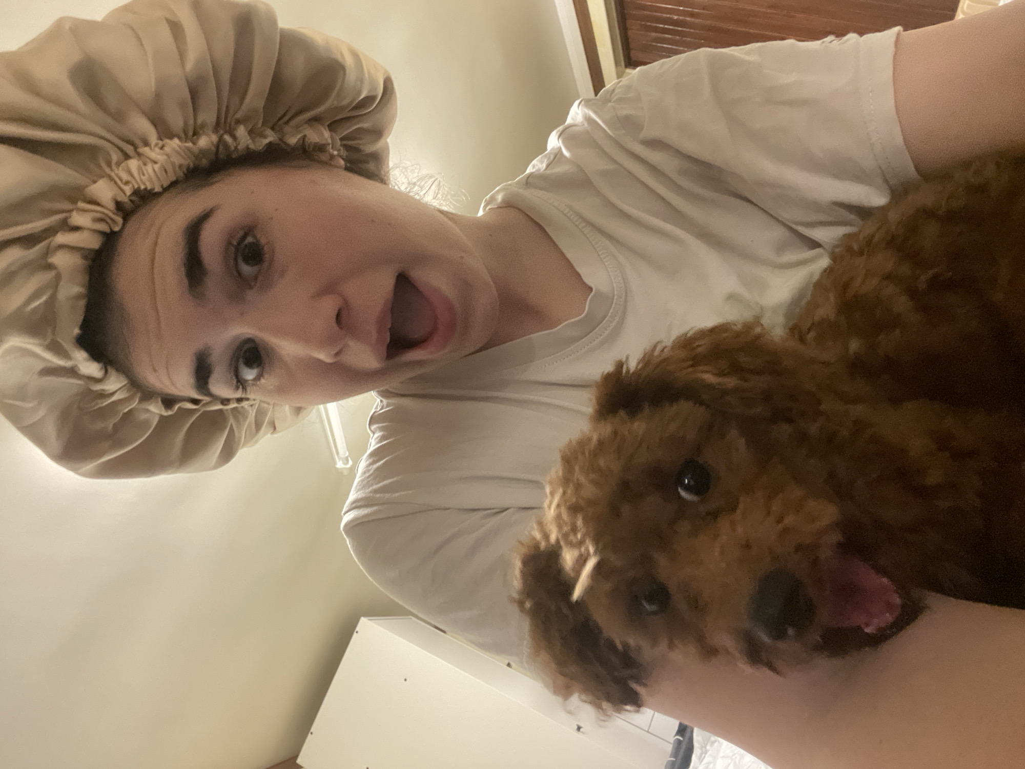 A selfie from a low angle shows a fair-skinned person with brown eyebrows and hair covered by a beige satin bonnet, mouth open in a wide expression. The person wears a white t-shirt and cuddles a brown dog with curly hair, whose mouth is also open. The background includes a white cabinet, a partially visible headboard made of wood, and a neutral wall. There is no indication of the city of Sofia, Bulgaria, or any other specific location.