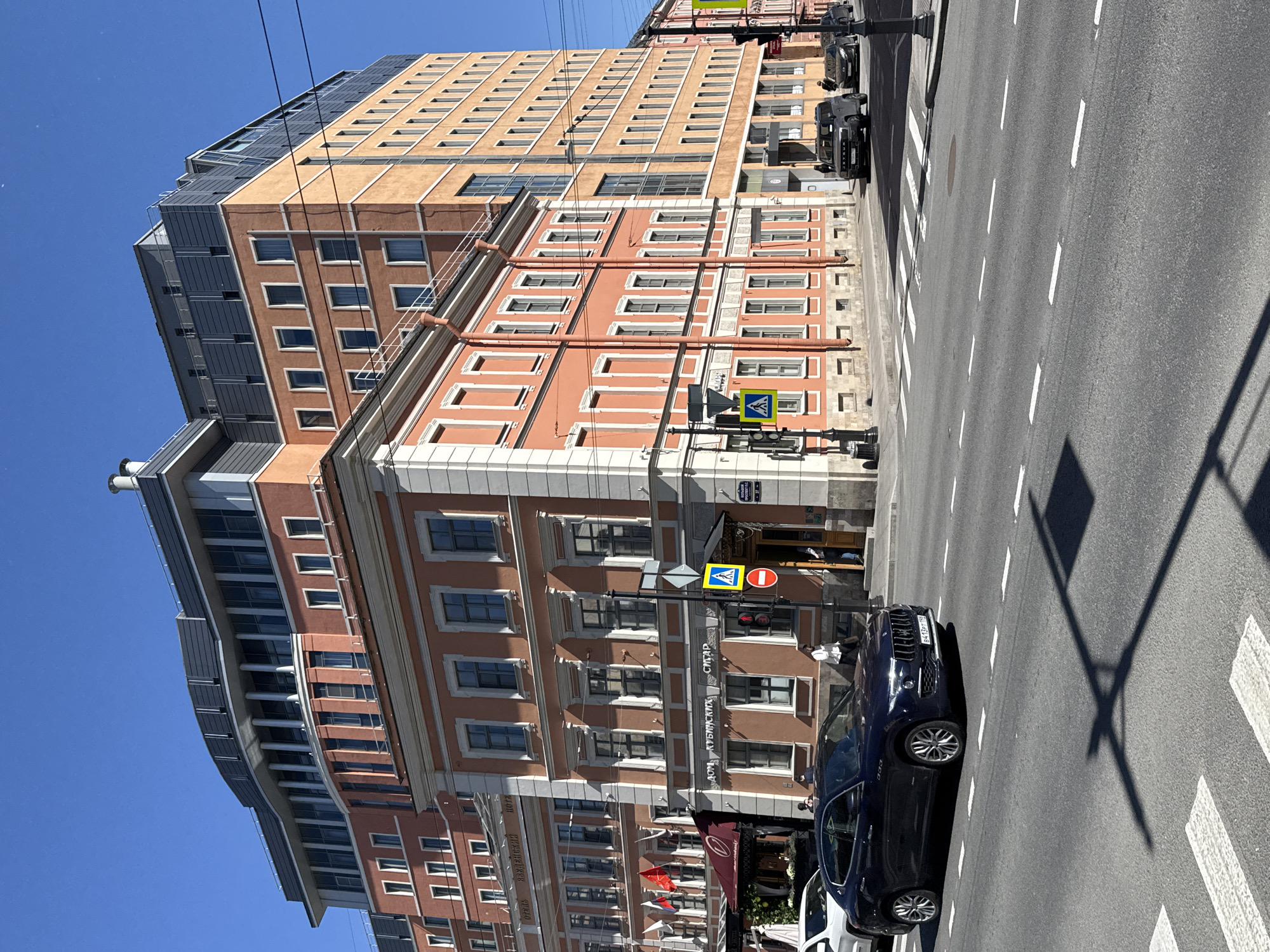 A sunny daytime, eye-level, exterior shot captures a street scene in an urban environment. The focal point is a multi-story building with an orange facade, featuring numerous windows and decorative white trim. Behind this building, a taller, modern building with a light orange facade and a gray, stepped roofline is visible. The street in the foreground has a crosswalk and is occupied by several vehicles. On the right side of the frame, a dark blue sedan is parked on the street. In the middle ground, a dark blue SUV is also parked. A white SUV is visible on the left side of the frame. The sky is clear and blue. Several traffic lights and street signs are present at an intersection. Pedestrians are seen walking on the sidewalk in front of the orange building.